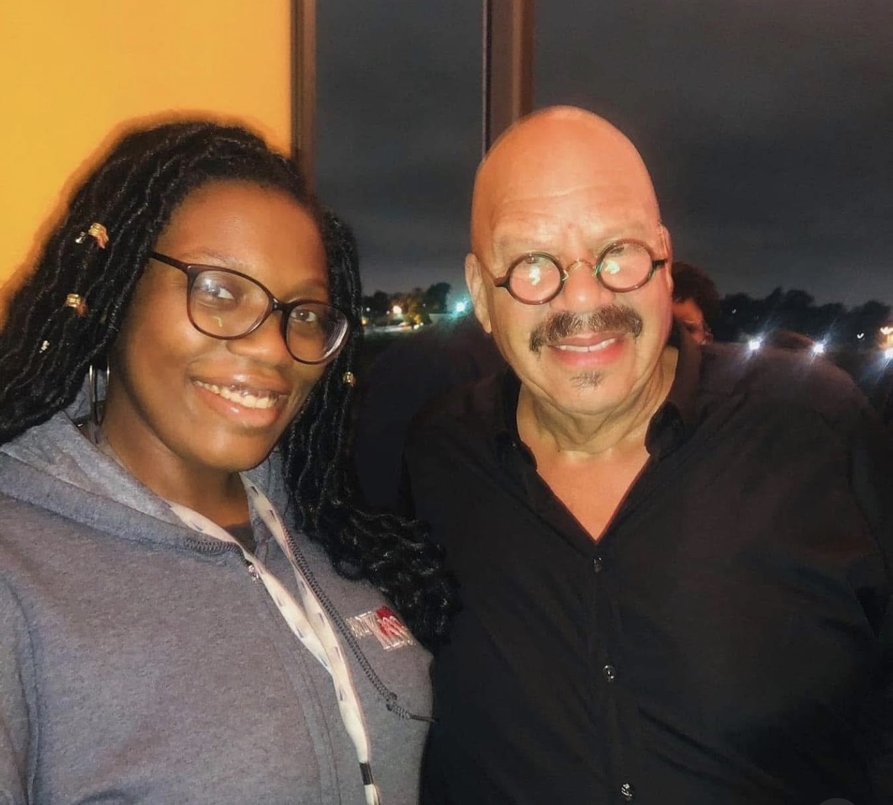 Two people smiling, one with glasses and dreadlocks, the other with glasses and a mustache, standing outdoors at night.