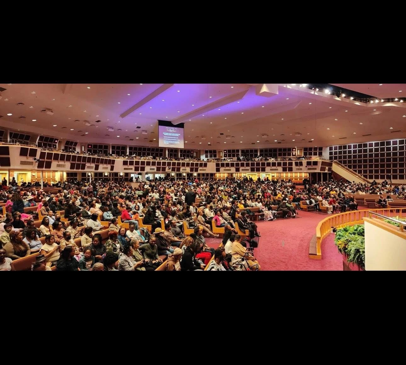 Large audience seated in a spacious auditorium with a stage and a large screen overhead.