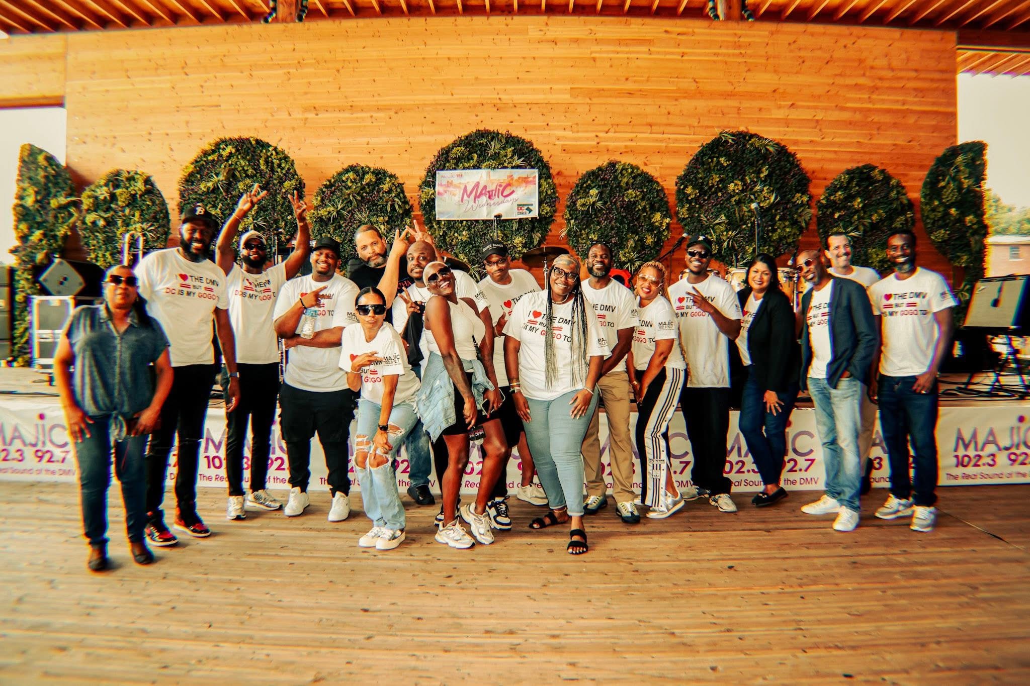 Group of people standing outdoors in front of a wooden building with trees, some holding signs, smiling, daytime.
