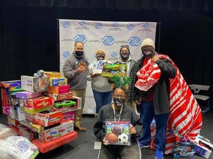 Five people wearing masks stand behind a table with toys and supplies, some holding items, in front of a backdrop.