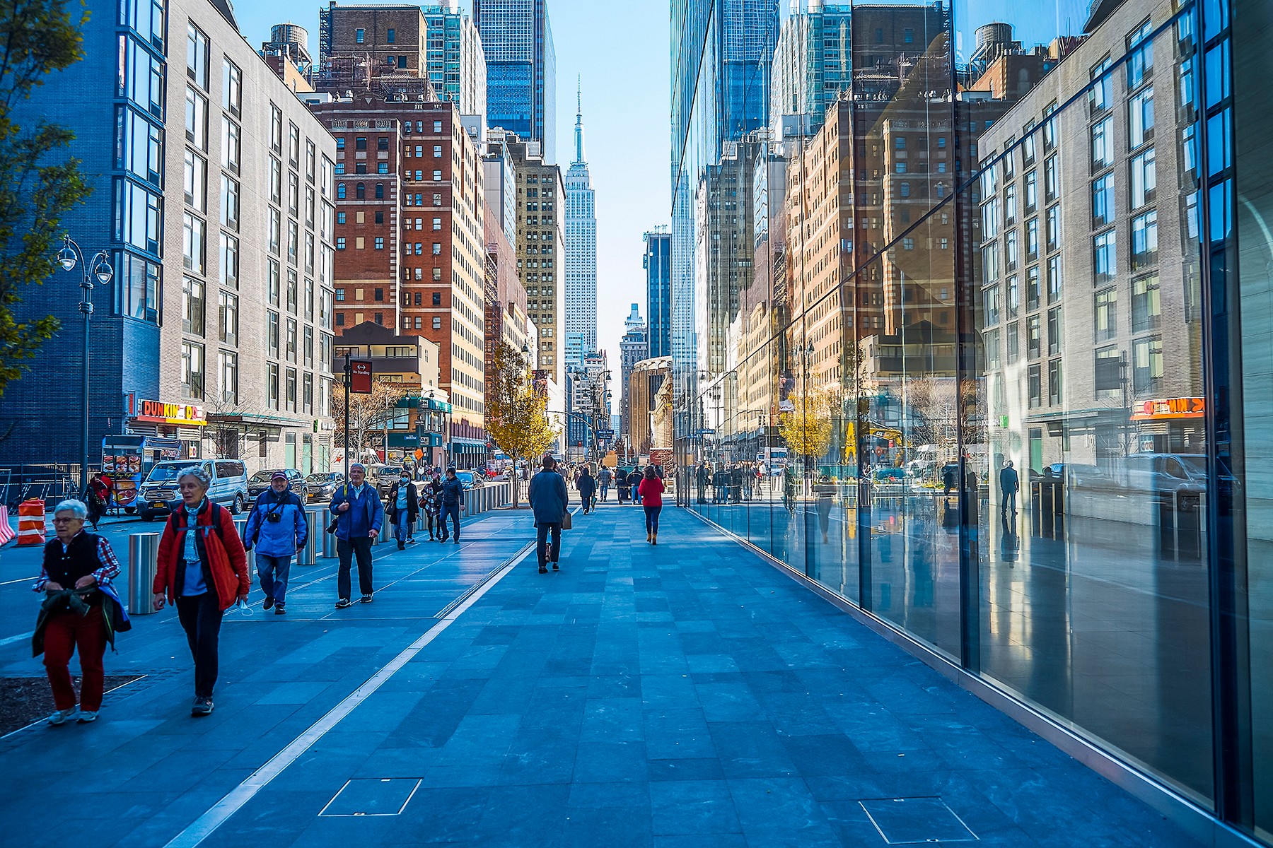 City street with pedestrians, tall buildings, and glass reflections, under a clear sky.