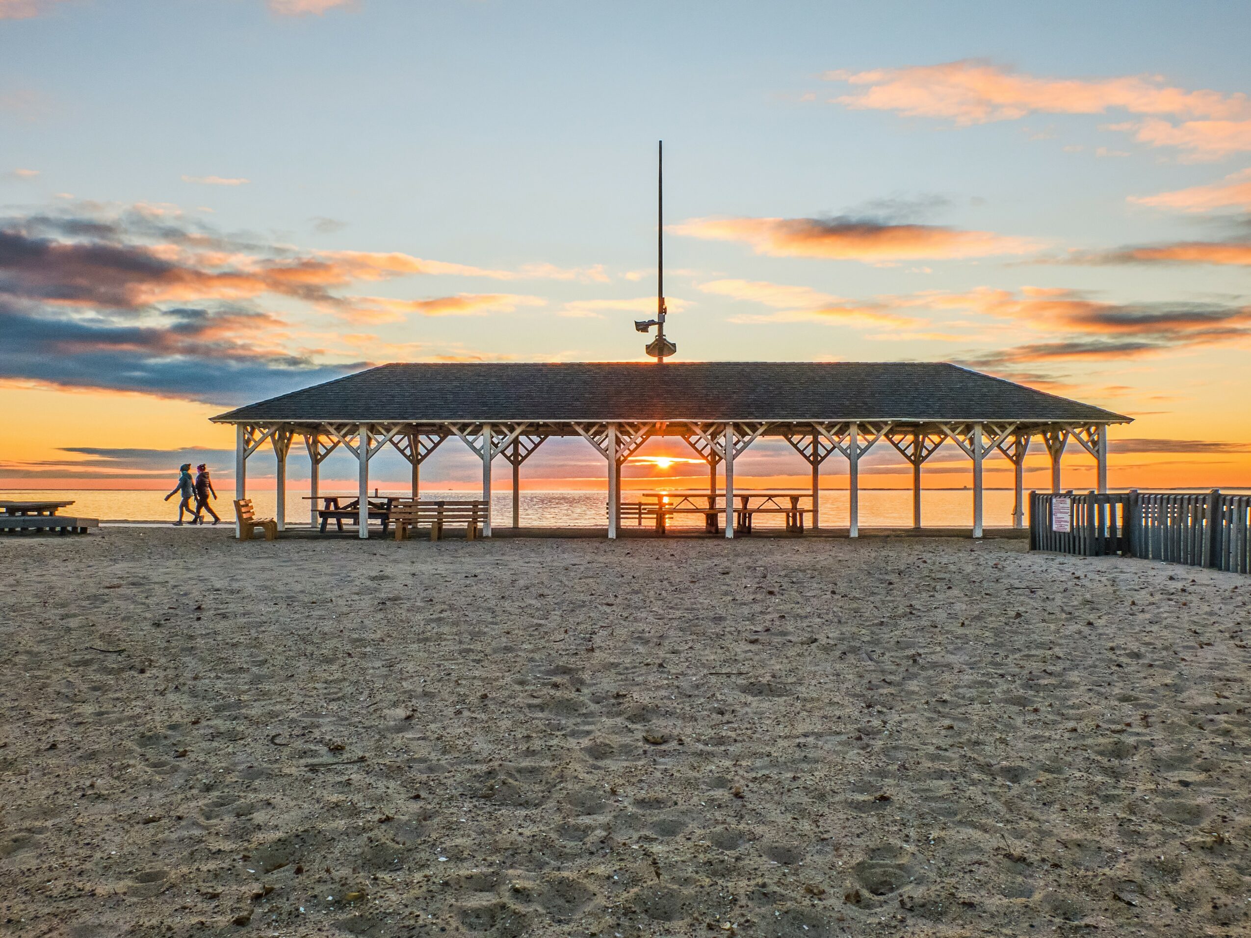 Beach pavilion with benches at sunset, sky with orange and pink clouds, sandy foreground, and water in the background.