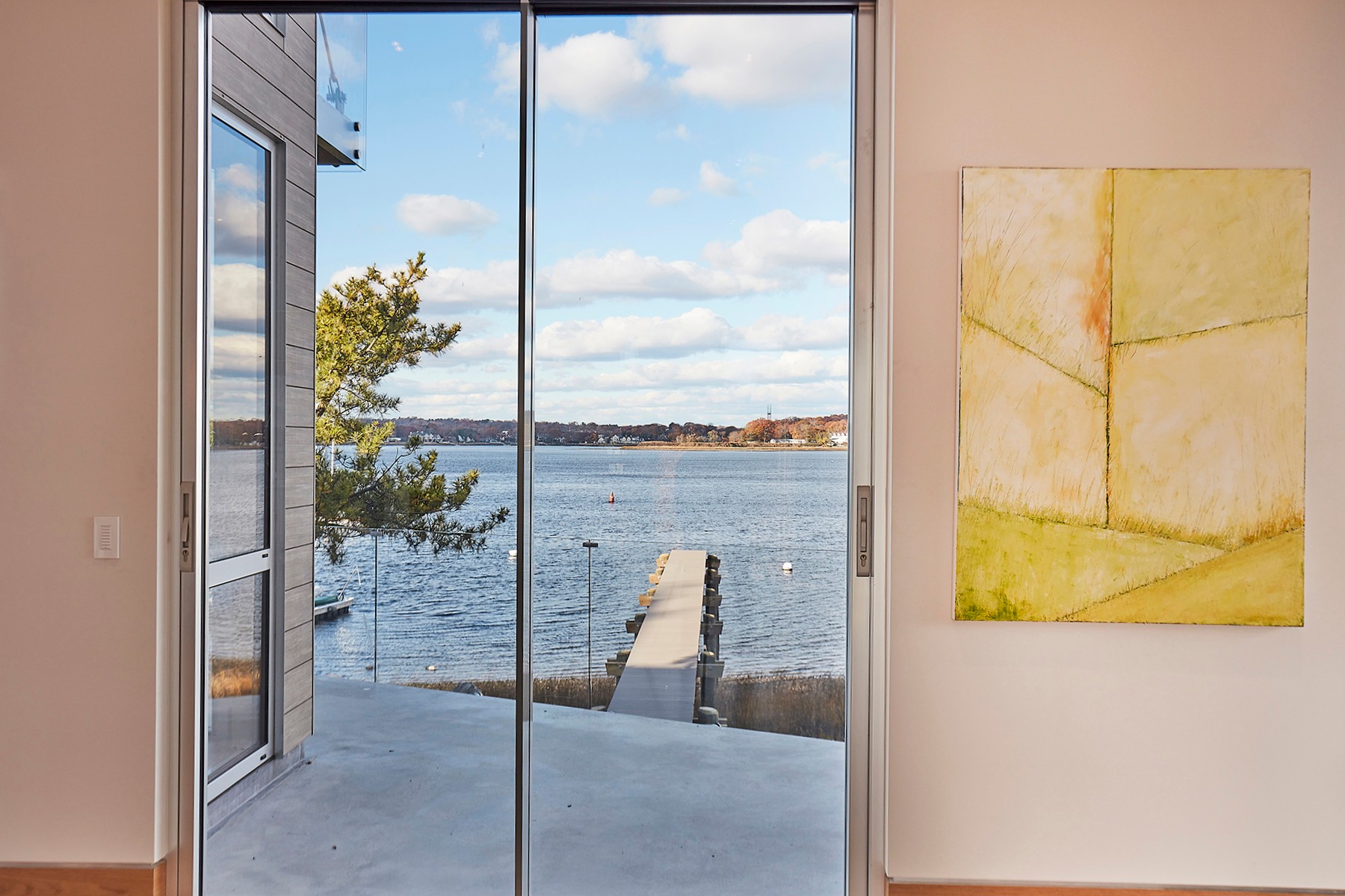 View through glass sliding doors showing water, a dock, and a tree outside, with a painting on the wall inside.