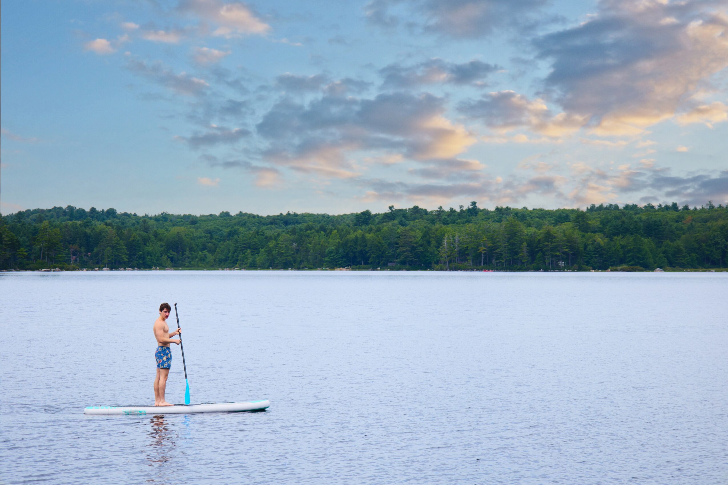 Person paddleboarding on a calm lake with a forested shoreline and partly cloudy sky in the background.