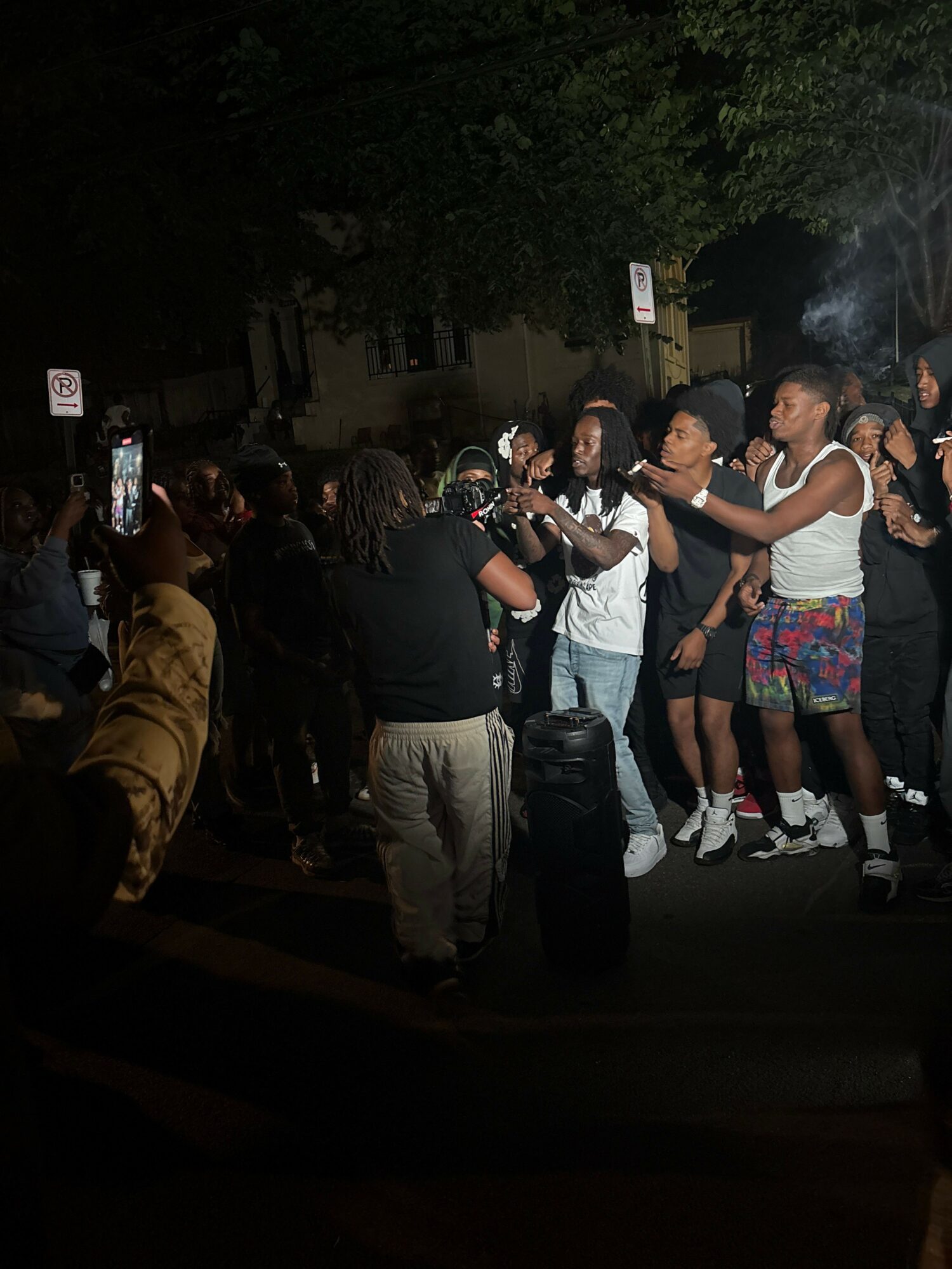 Group of people gathered outdoors at night, some holding drinks, engaging in conversation, with trees and lights in background.