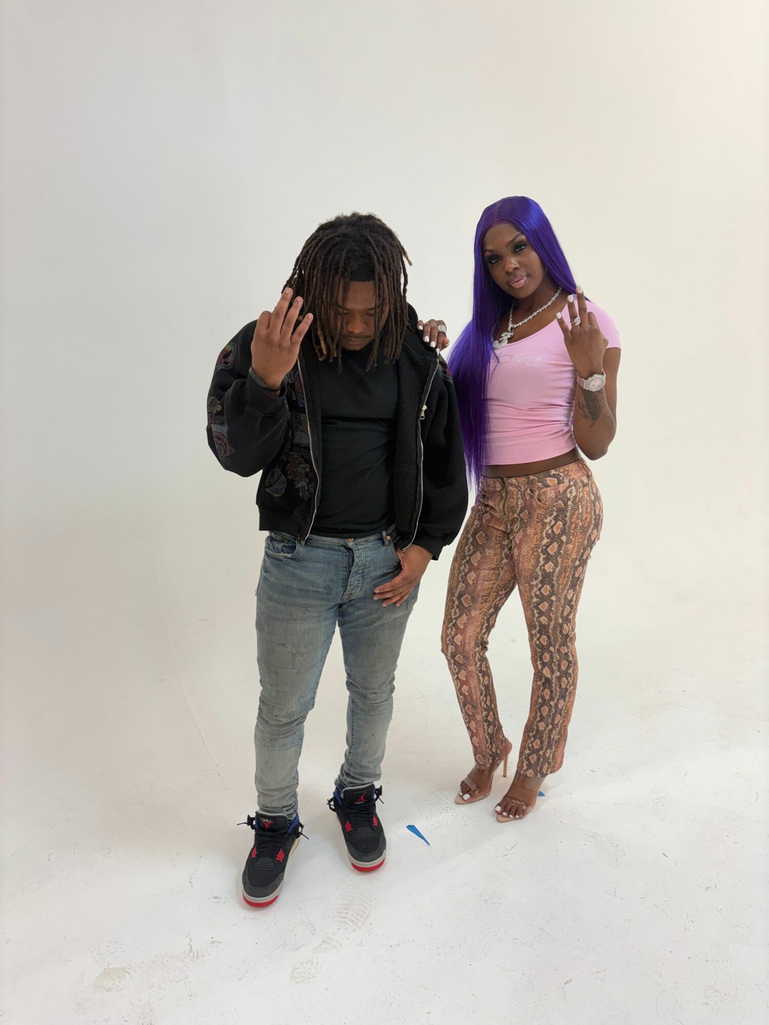 Two women standing against a plain white background, one in a pink top and patterned pants, the other in a black jacket and jeans.