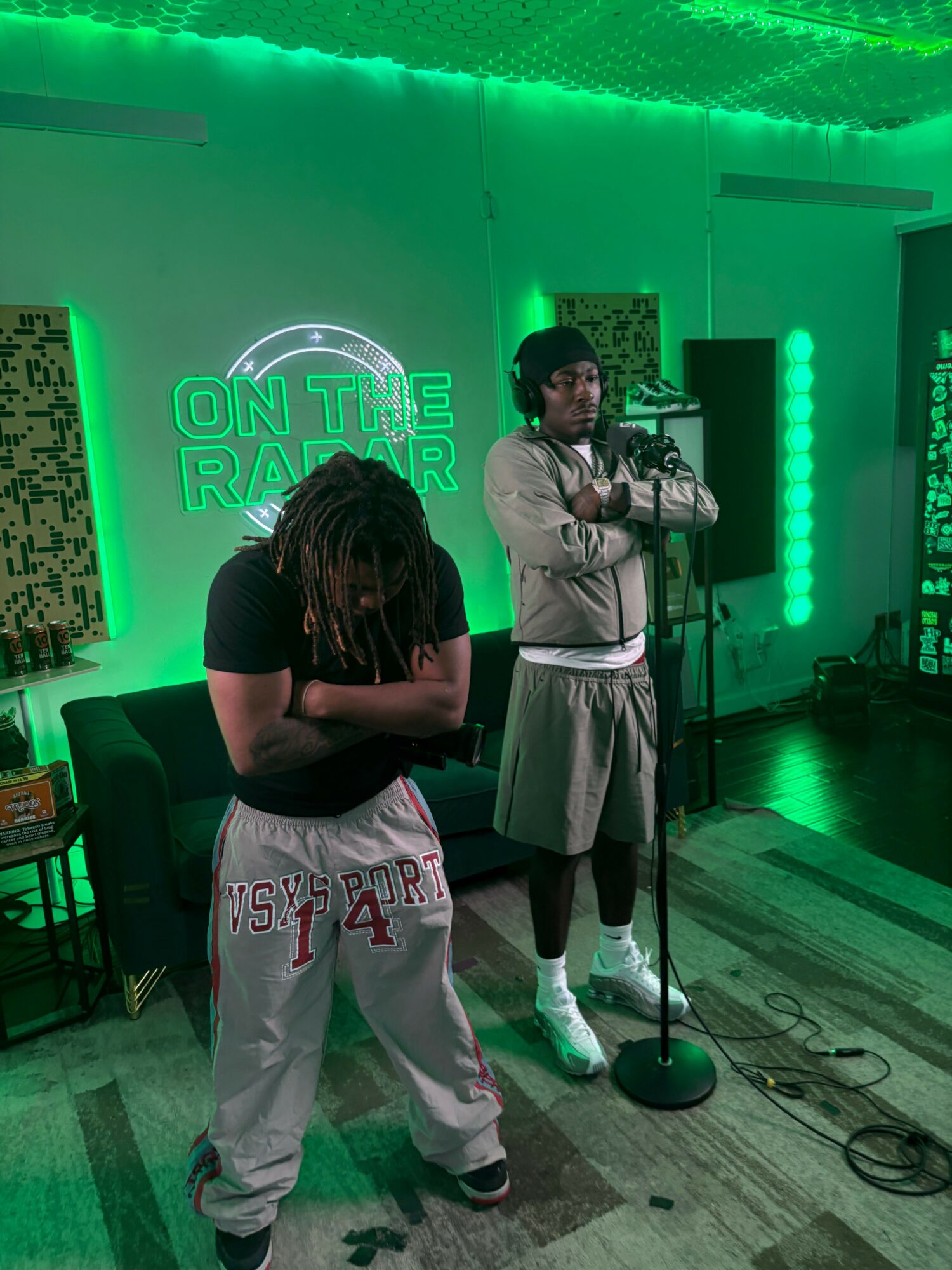 Two people stand in front of a green-lit wall with neon signs, one with dreadlocks and the other with short hair, in a room with wooden flooring.