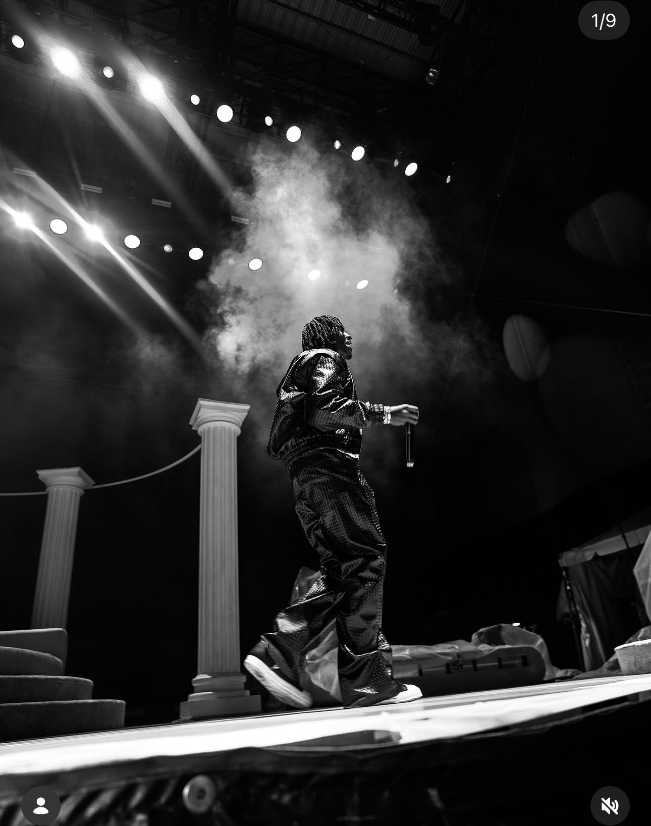 Performer on stage with smoke and bright lights, holding a microphone, wearing dark clothing, in a concert setting.