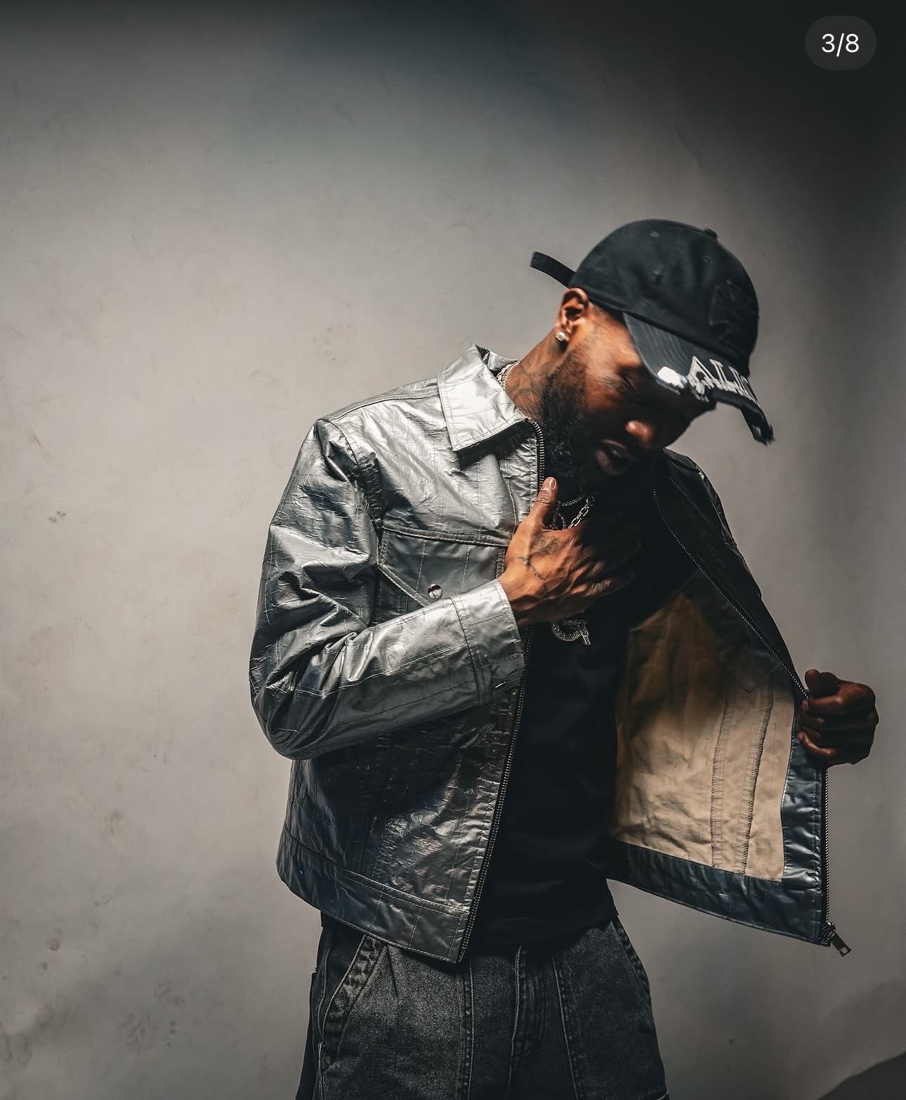Man wearing a black cap and metallic jacket, looking down and holding open his jacket, against a plain background.