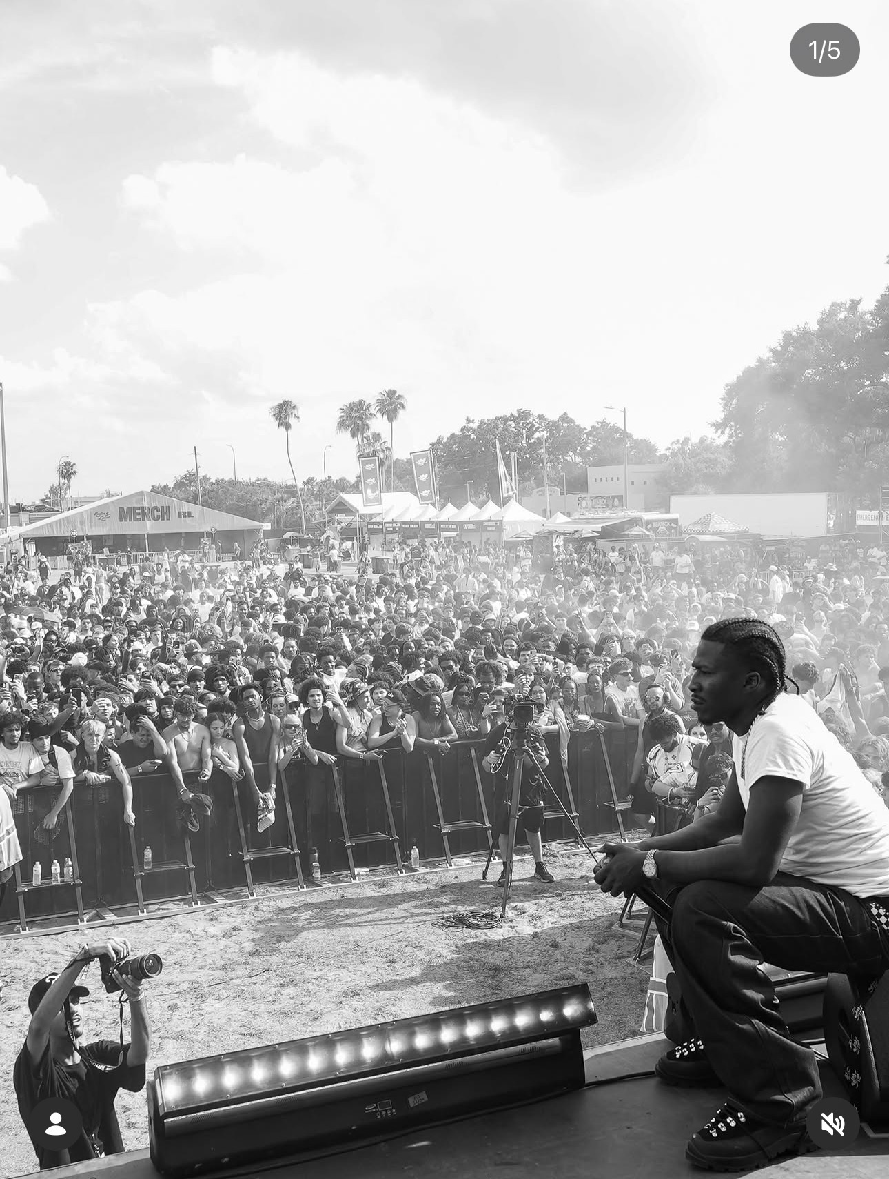 Person sitting on stage with a large crowd in the background, some trees and buildings visible, outdoor event, sunny day.