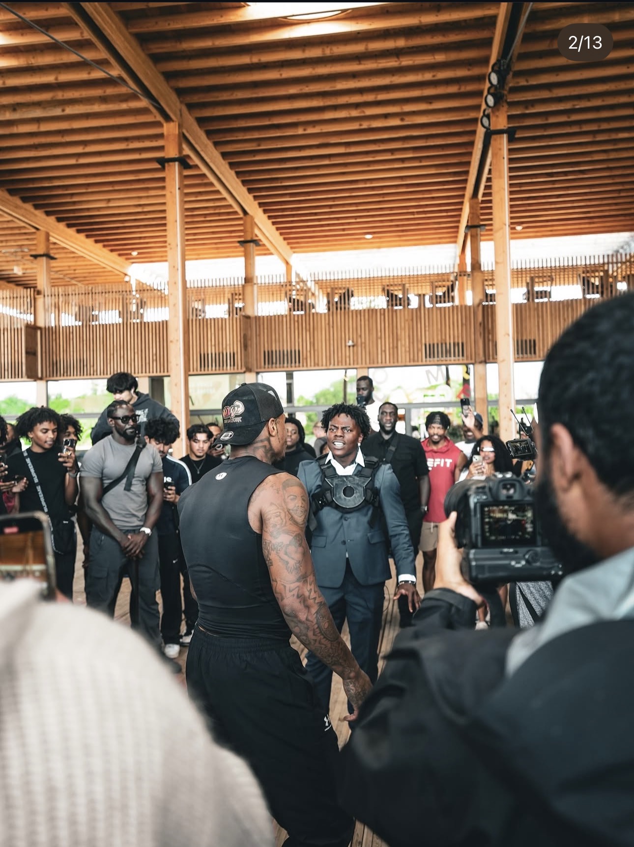 Group of people gathered outdoors under a wooden roof, some taking photos or videos, with a man in a black sleeveless shirt in the center.