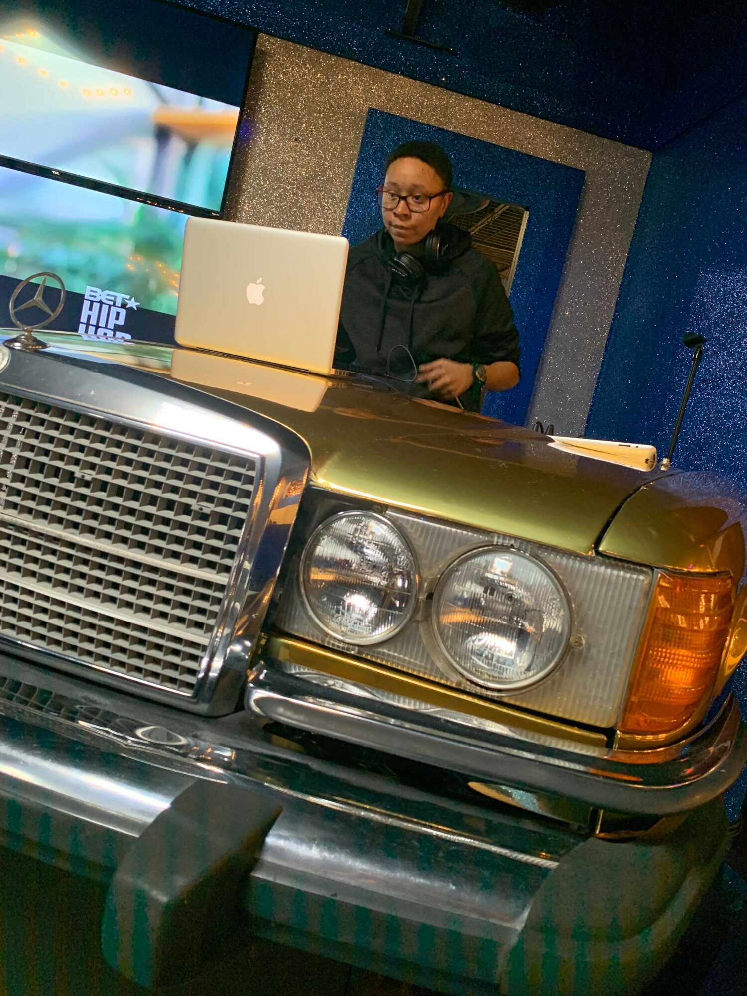 Person standing behind a vintage car with a laptop on the hood, in an indoor setting.