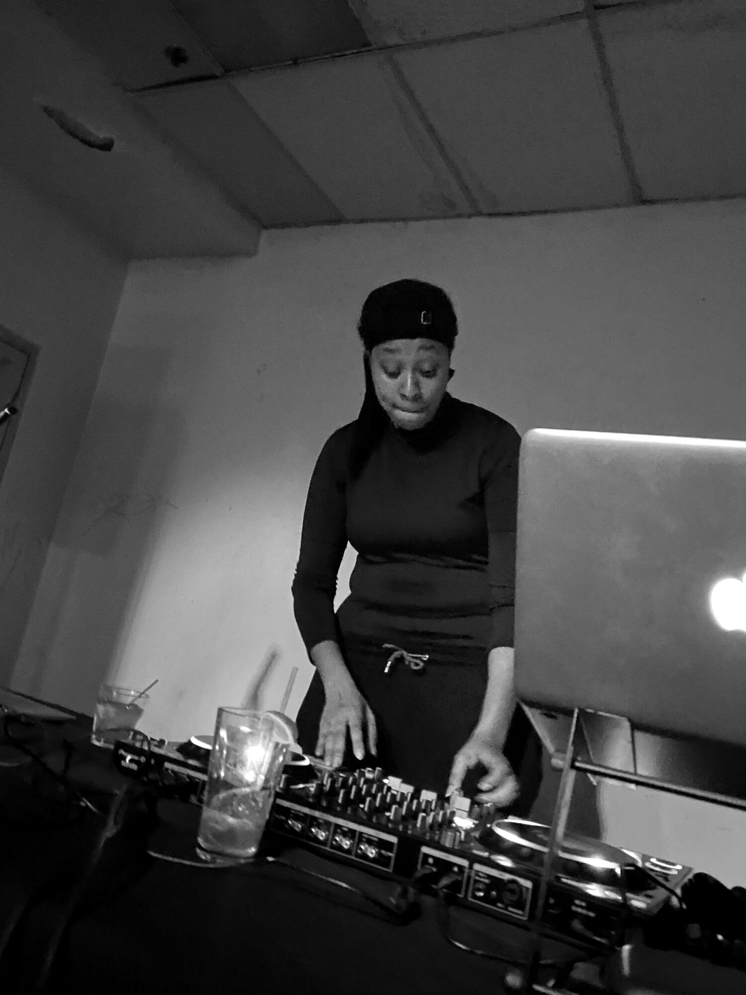 Person DJing with equipment and a laptop in a room, black and white photo.