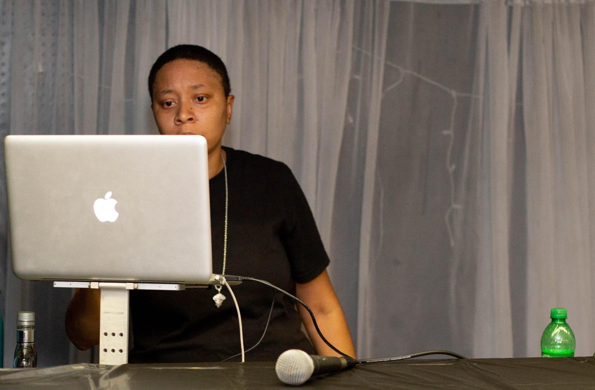 Person standing behind a table with a laptop, microphone, and a green bottle, in front of curtains.