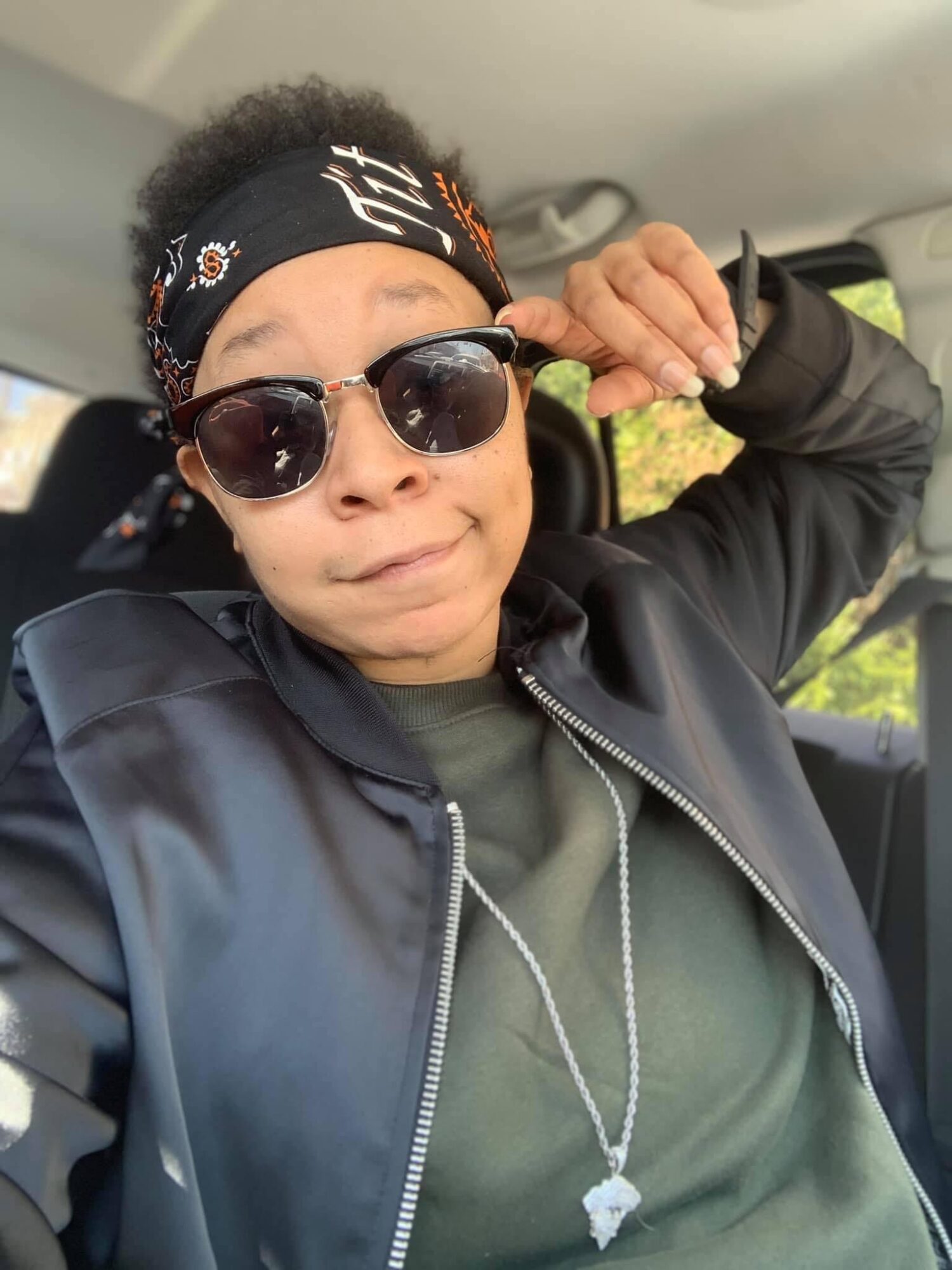 Person with sunglasses and headband, wearing a black jacket, sitting in a car, making a fist gesture.