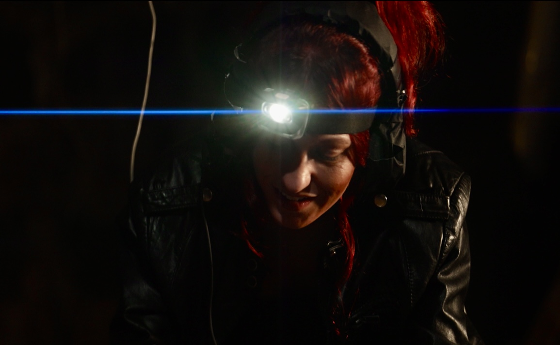 Person wearing a headlamp with a bright light, dark background, and red hair, looking downward.