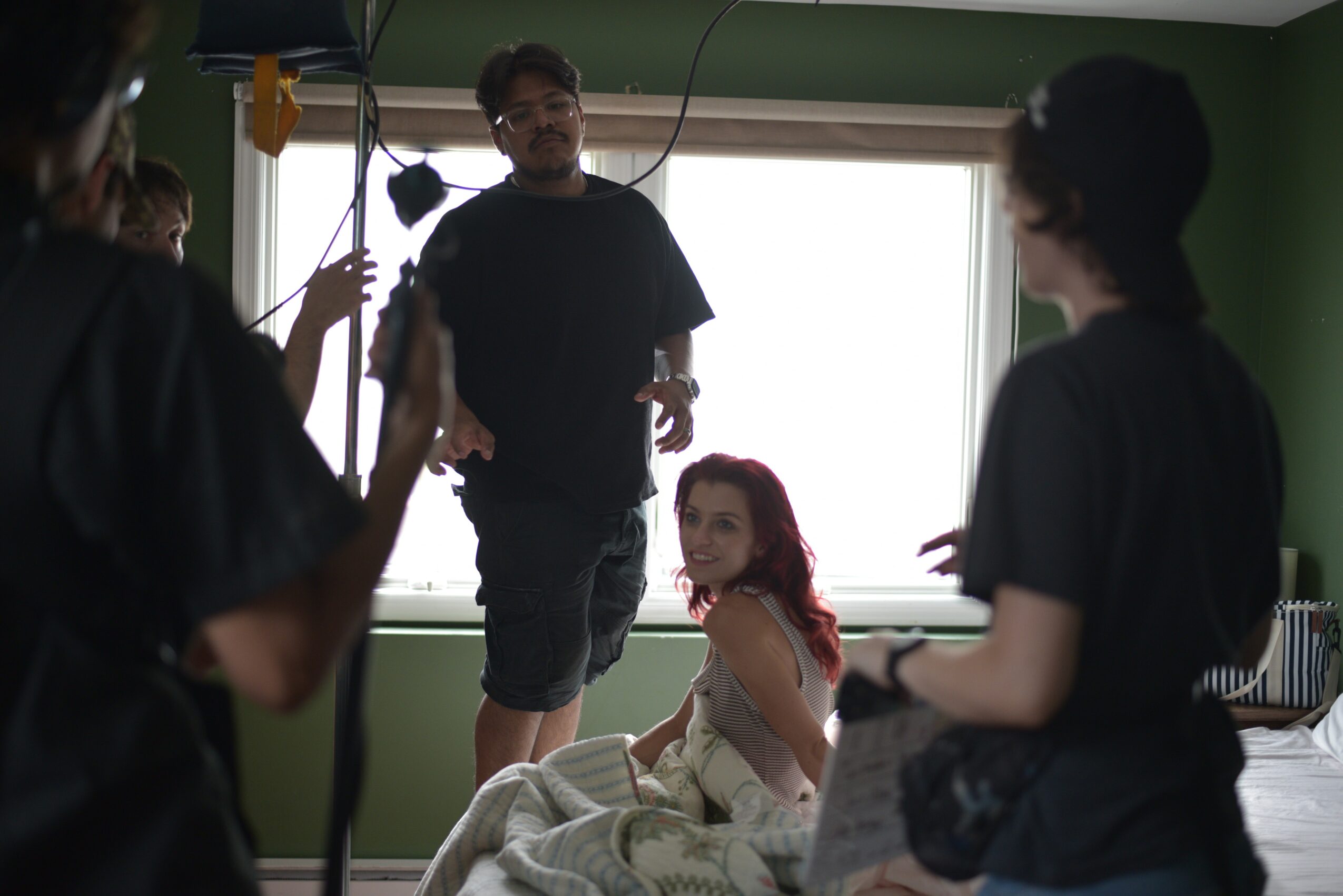 Group of people in a room with green walls and a large window, one person sitting on a bed, others standing around.