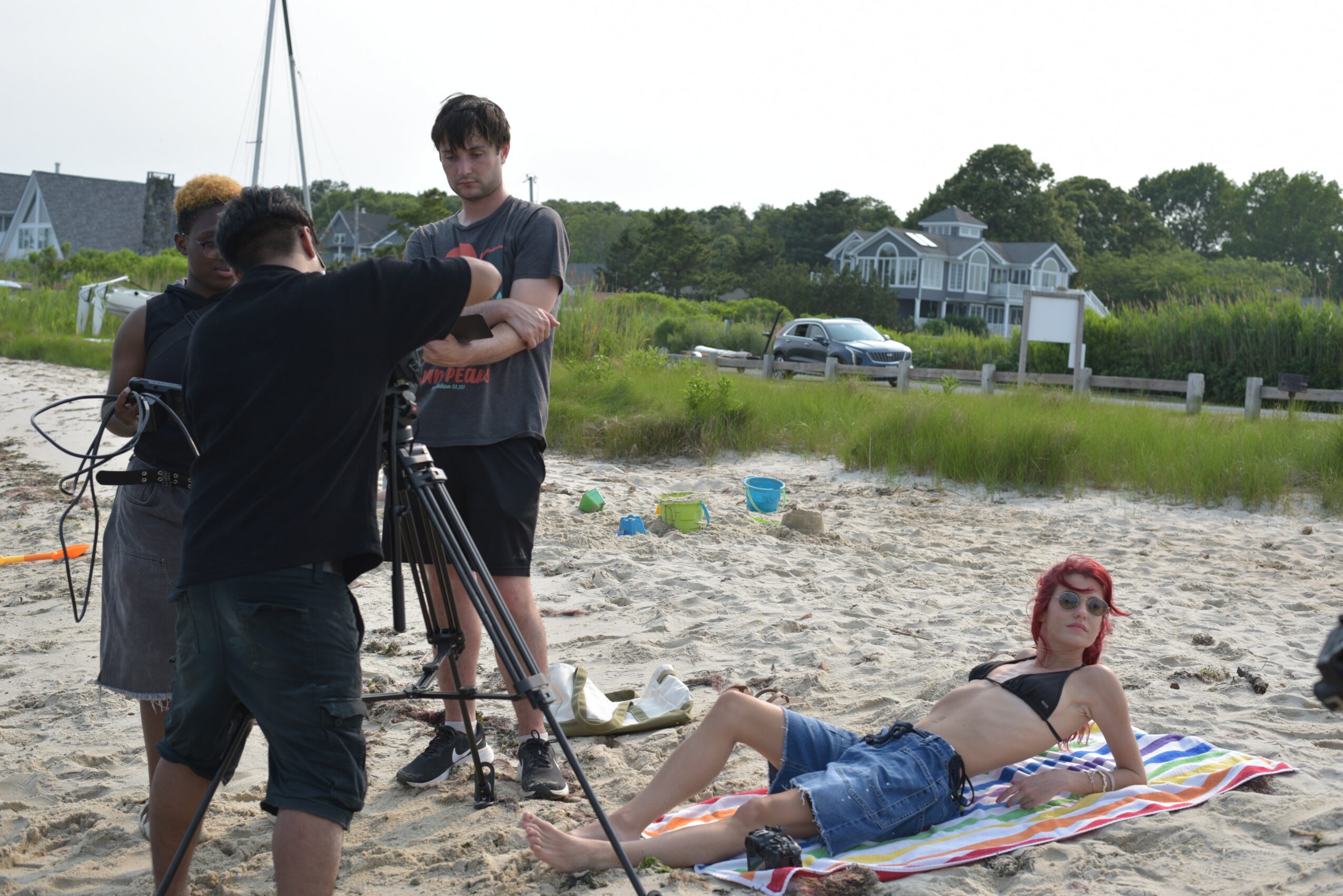 Group of people setting up equipment on a sandy beach with a woman lying on a towel, houses, and greenery in background.