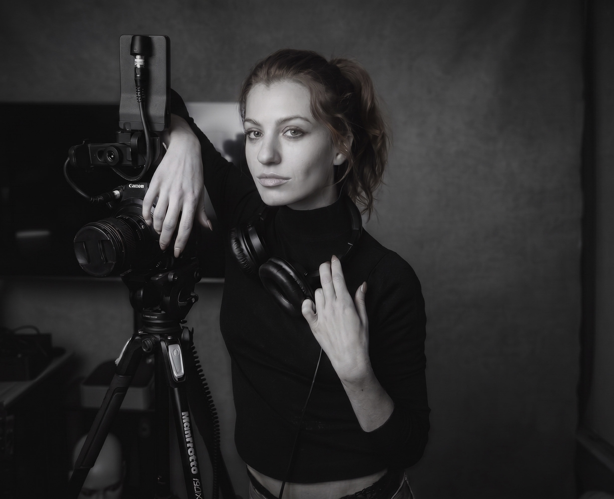 Woman with headphones and camera equipment, looking at the camera, in a dark studio setting.