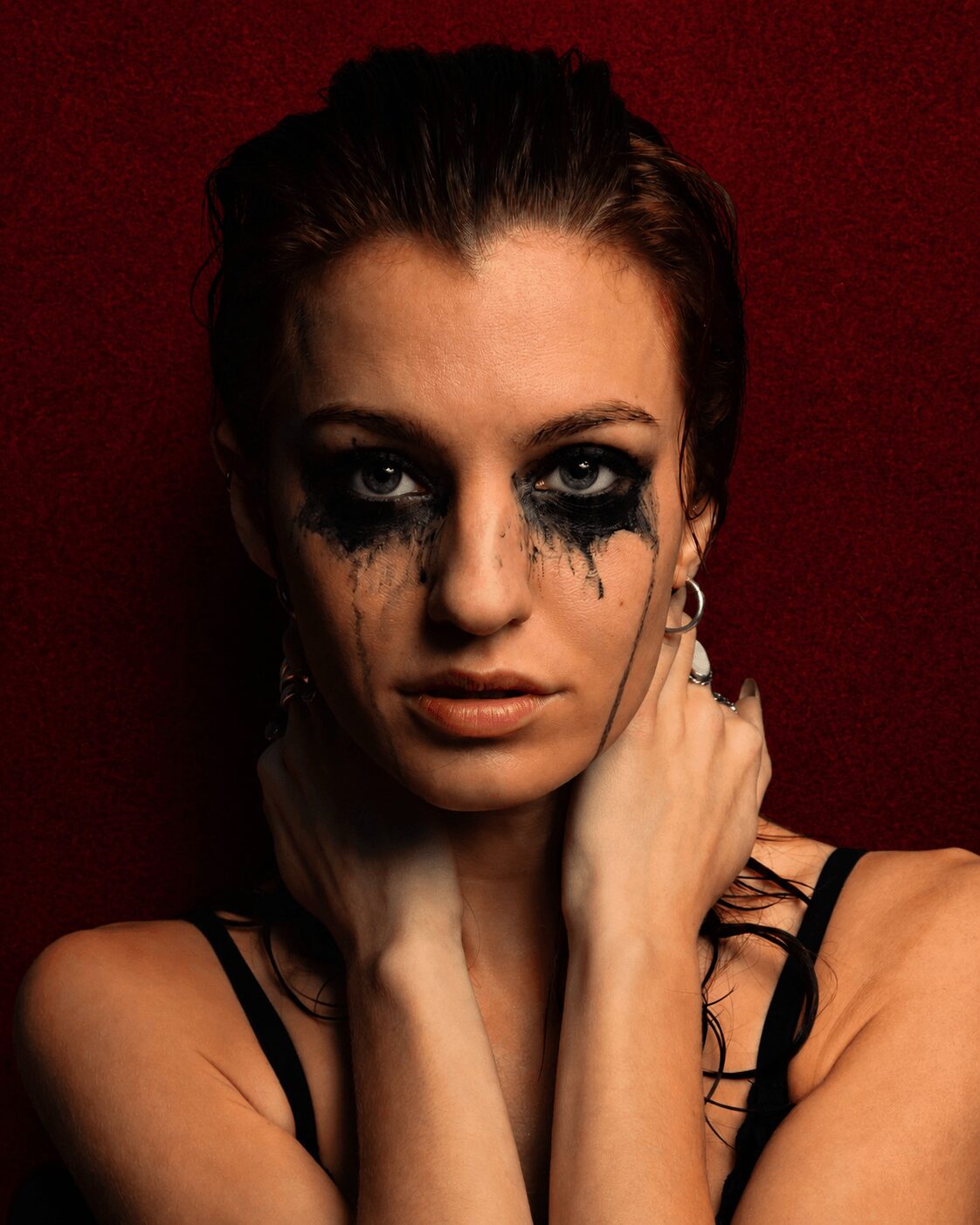 Woman with dark makeup around eyes, dark smudges on face, holding her neck, against a dark red background.