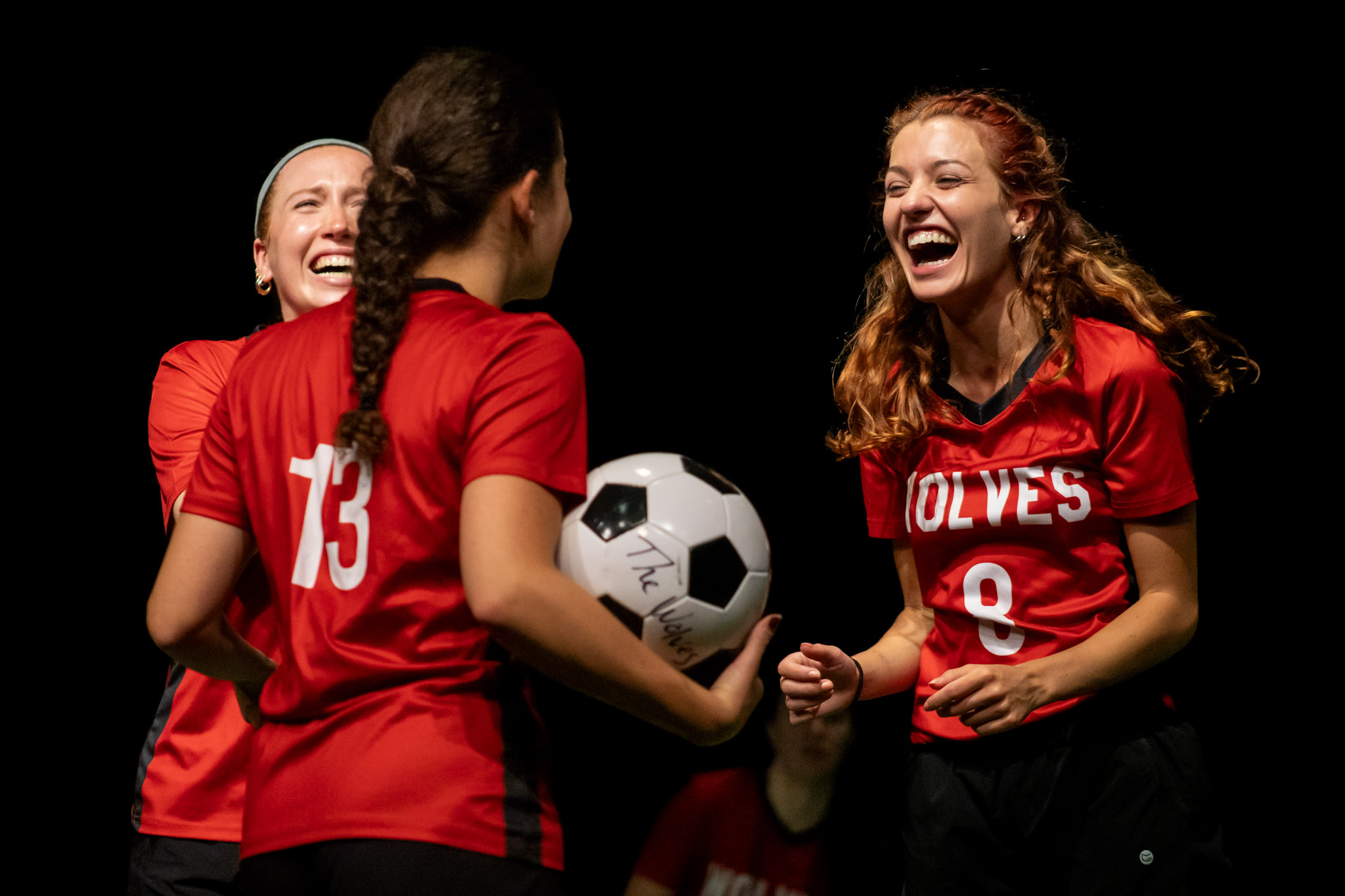 Three women in red soccer jerseys smiling and celebrating, one holding a soccer ball, against a black background.