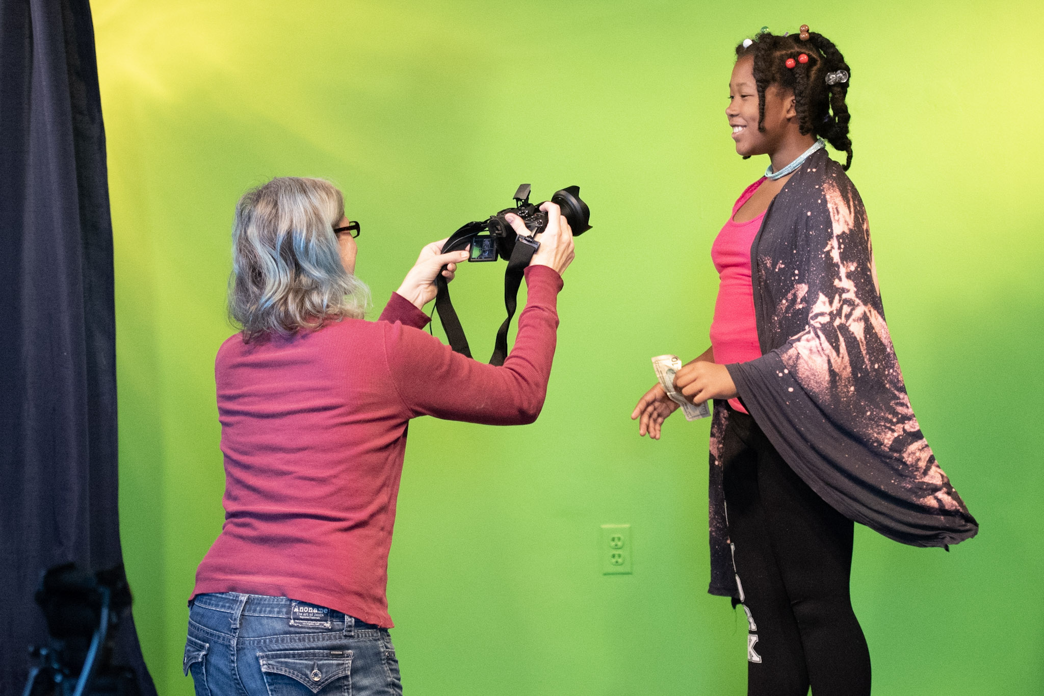 A woman with gray hair and glasses takes a photo of a young girl with braided hair on a green background.