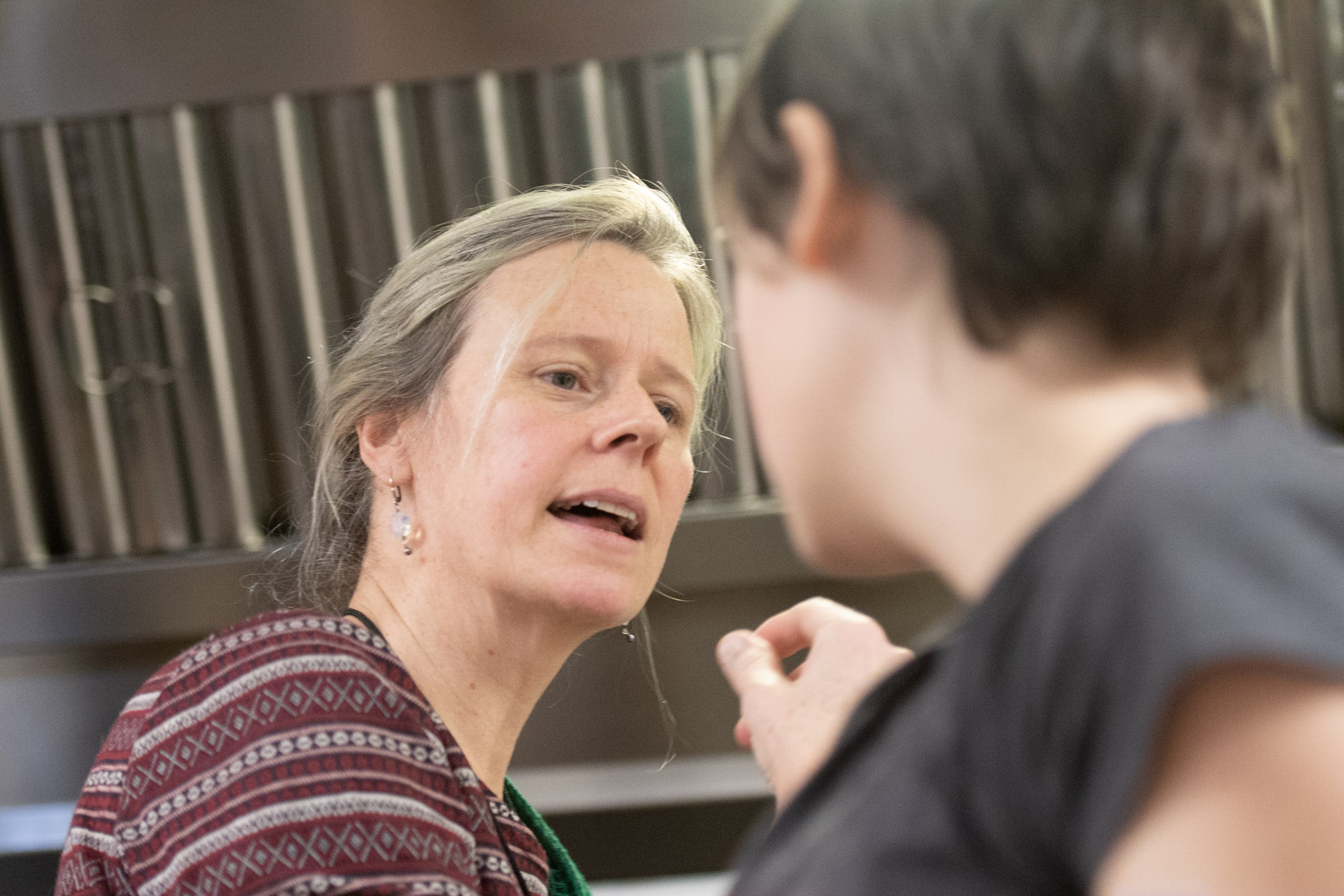 Older woman with gray hair speaking to a person with short dark hair in a kitchen setting.