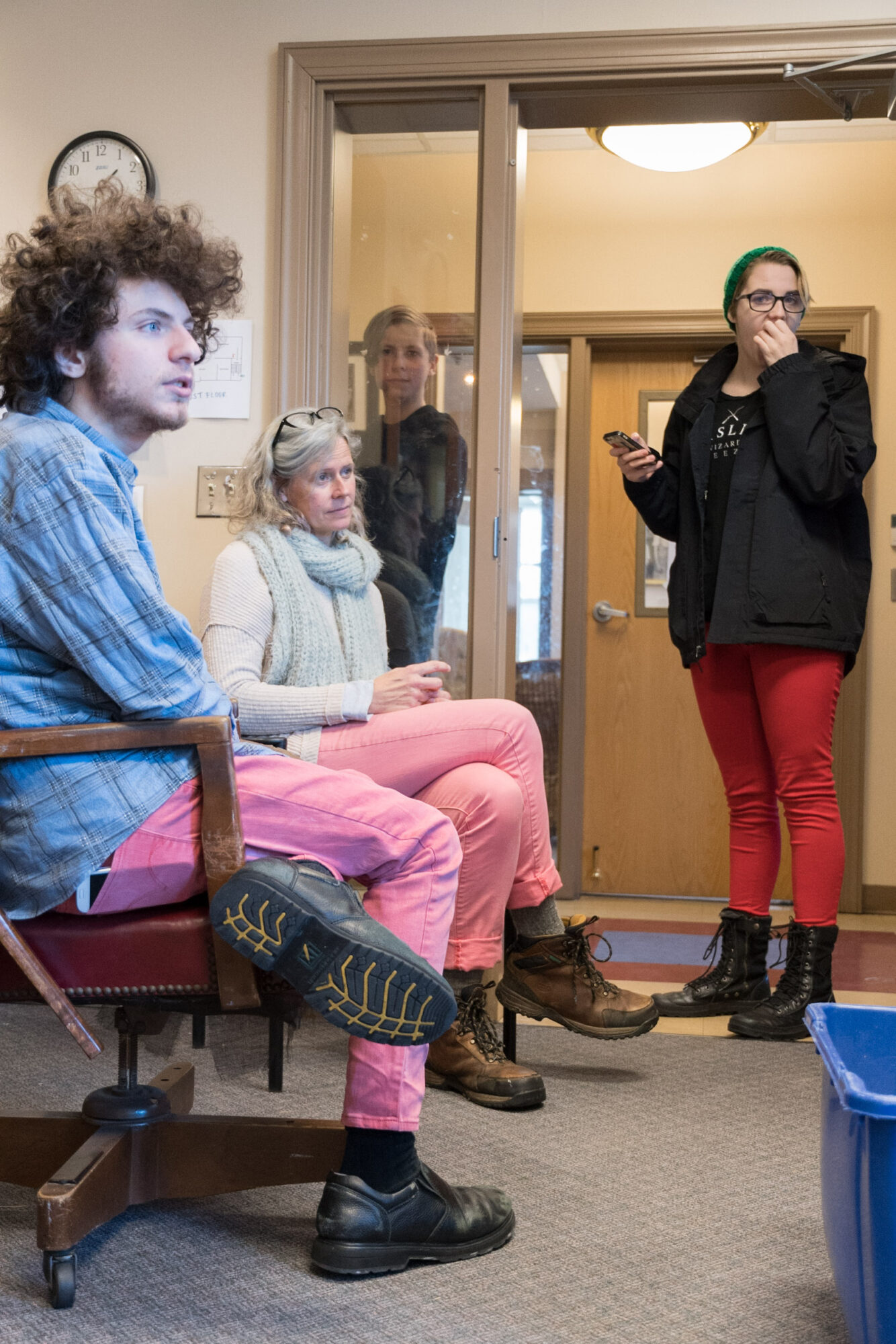 Two women and a young girl sit on a bench inside a room, with a woman standing nearby holding a phone, in front of a glass door.