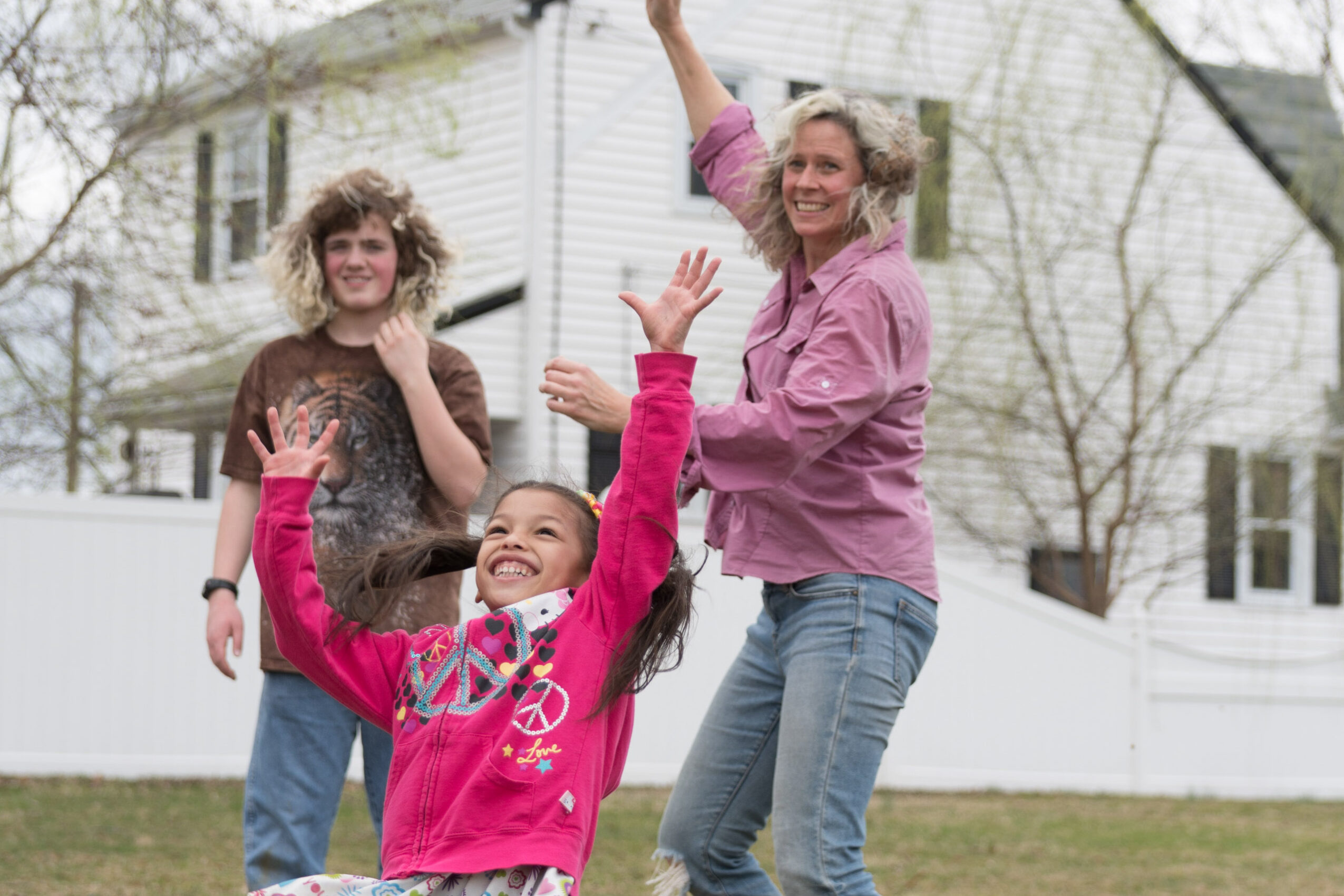 Three women outdoors, smiling and waving, with trees and a white house in the background.