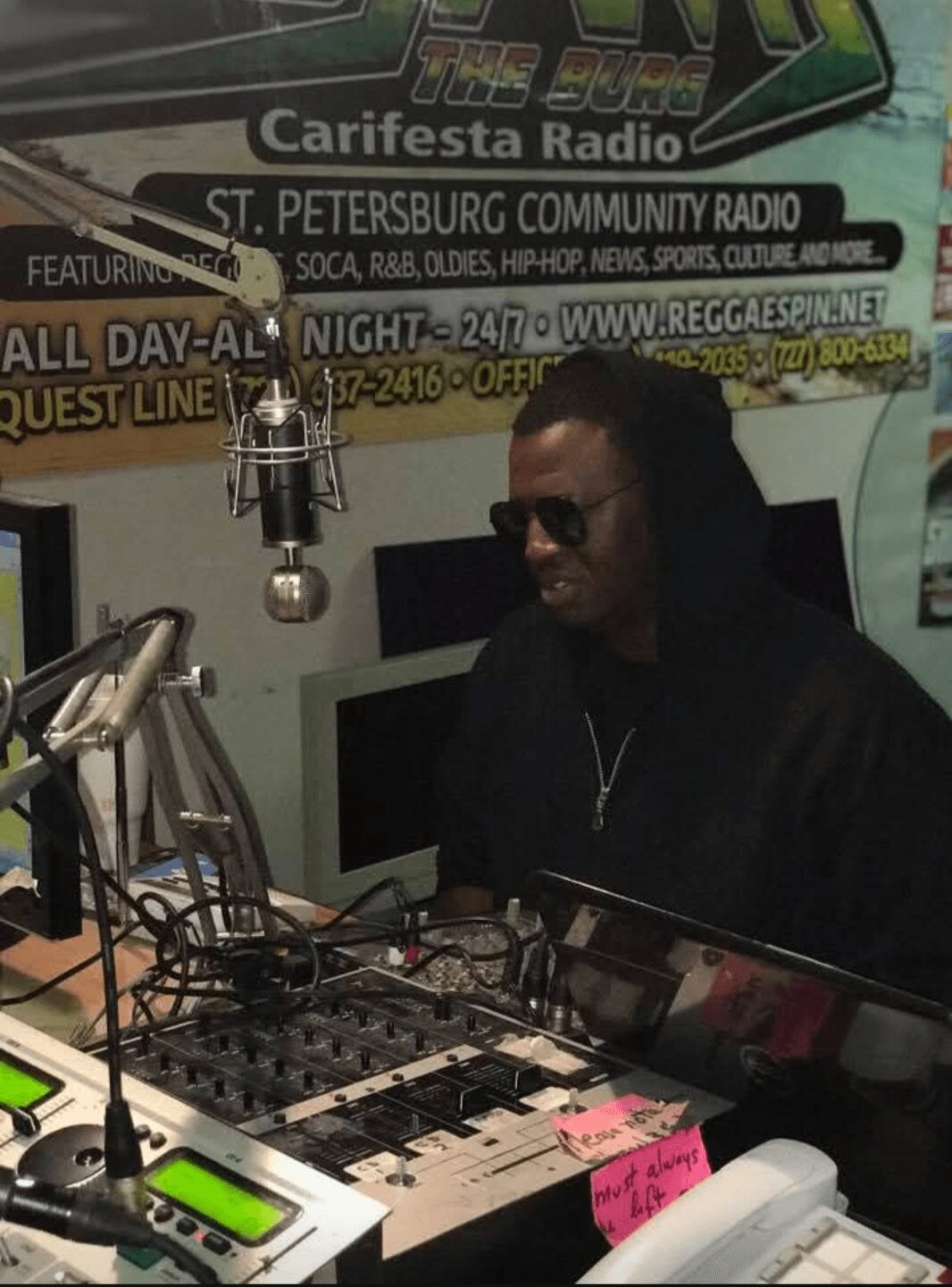 Person wearing sunglasses sitting in a radio studio with equipment and microphones, promotional posters on the wall behind.