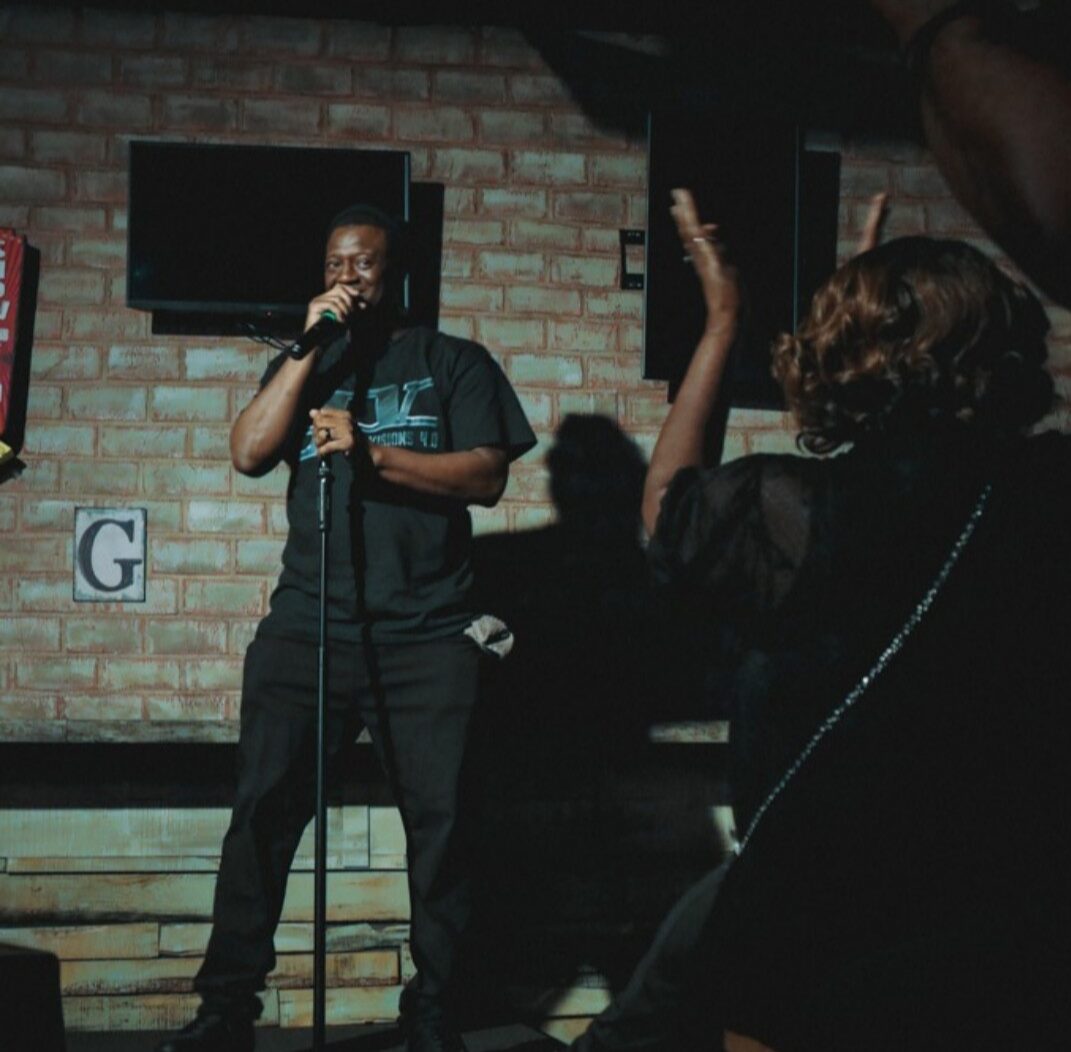 Person singing into microphone on stage with audience raising hand, brick wall background, TV screen, and letter G visible.