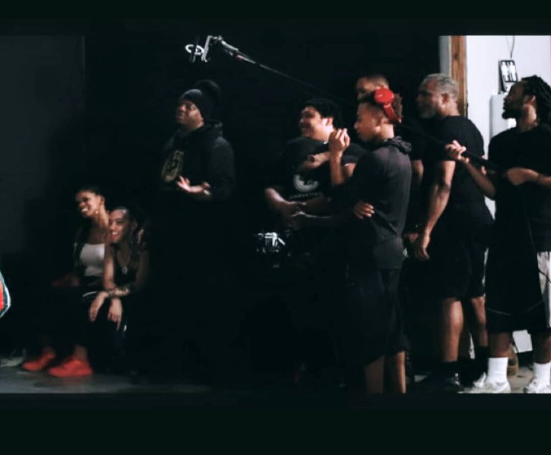 Group of people standing and sitting in a dark room, some wearing black clothing, with a microphone overhead.