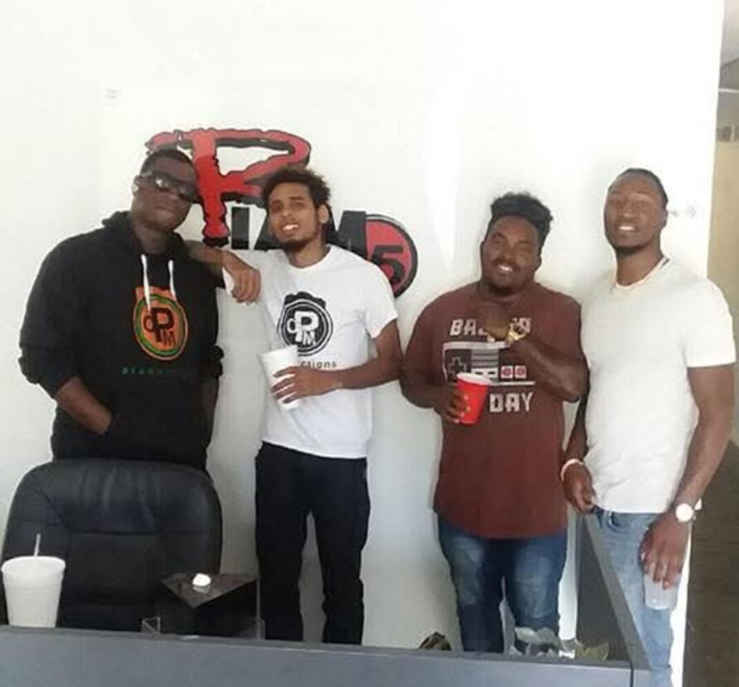Four men standing indoors, smiling, with a logo on the wall behind them, holding cups, in casual clothing.