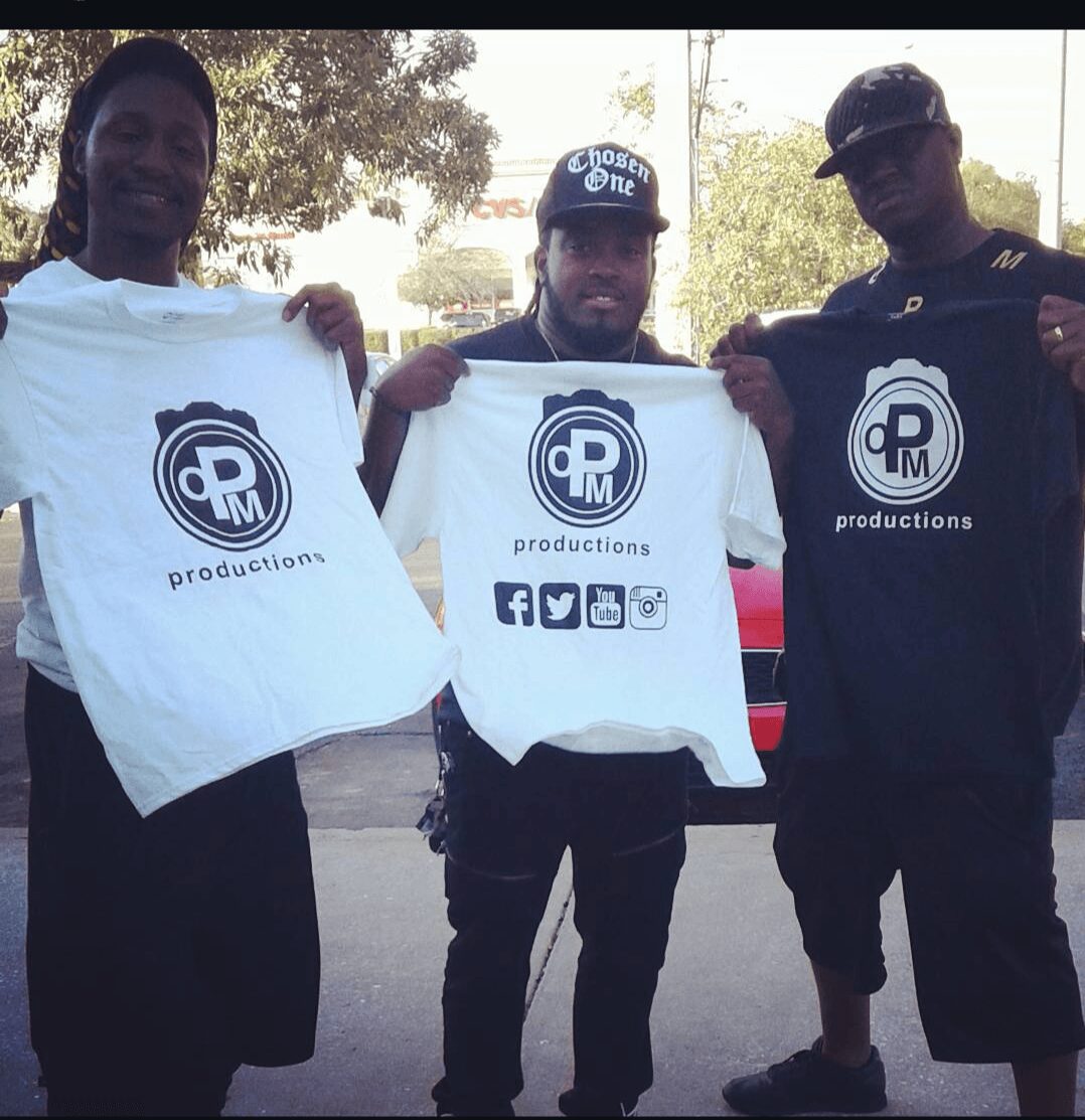 Three men holding white T-shirts with a logo and social media icons, outdoors with trees and cars in background.