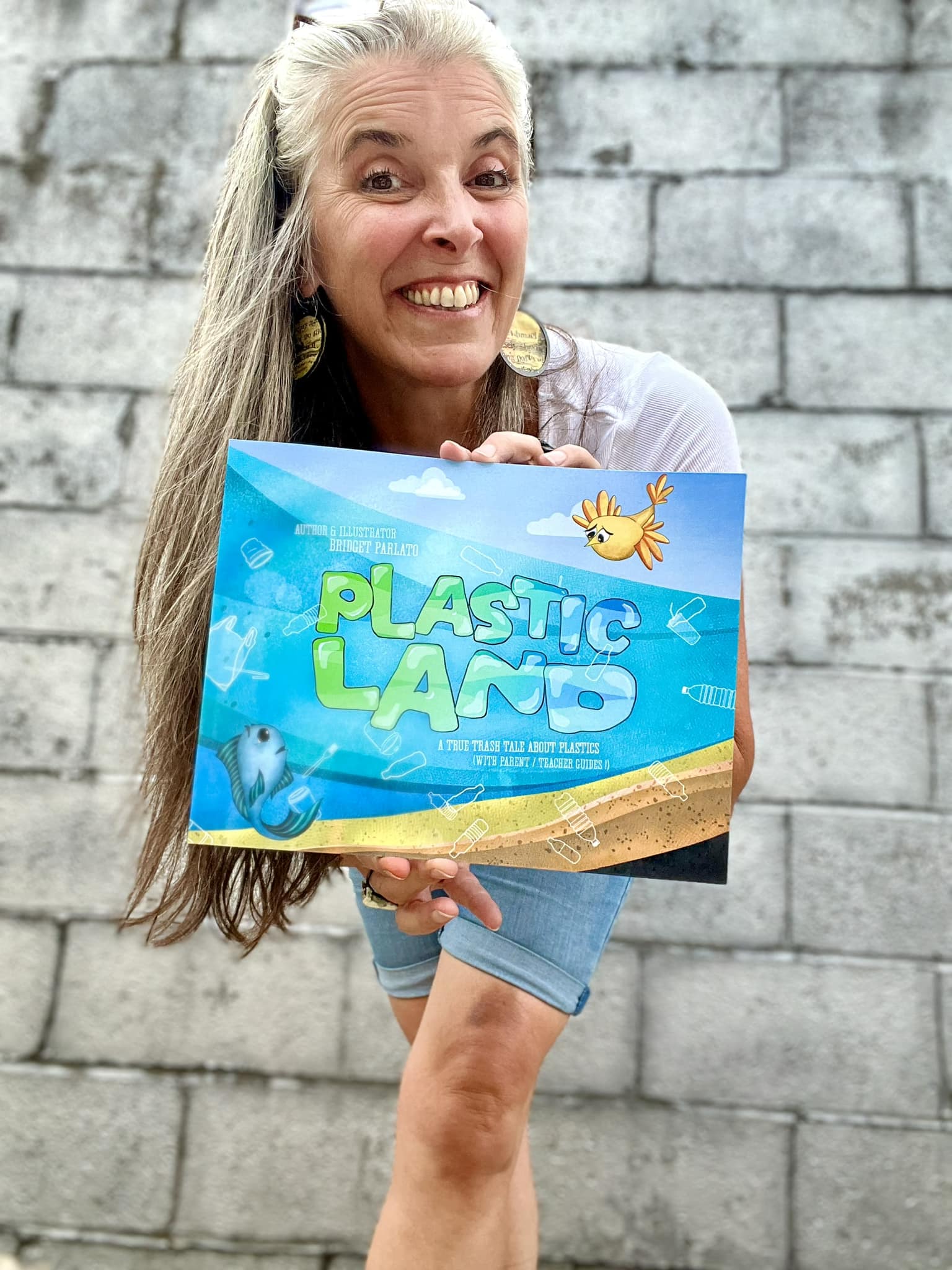 Smiling woman holding a colorful sign that says 'Plastic Land' in front of a gray brick wall.