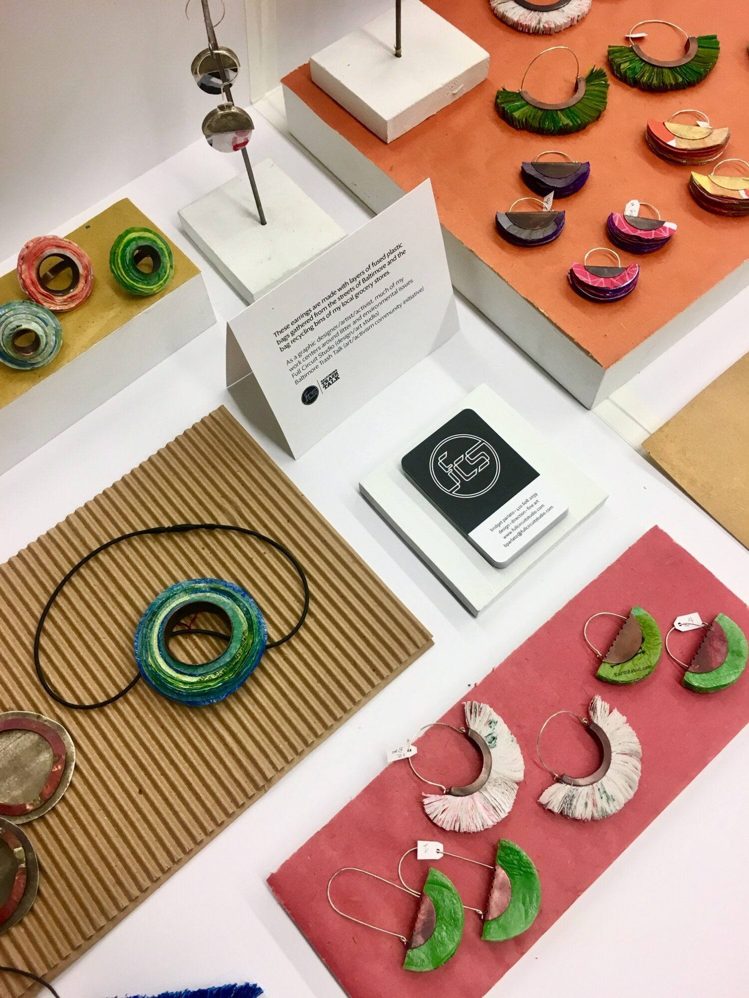 Display of colorful jewelry including necklaces and earrings on white and pink surfaces, with some pieces on textured backgrounds.