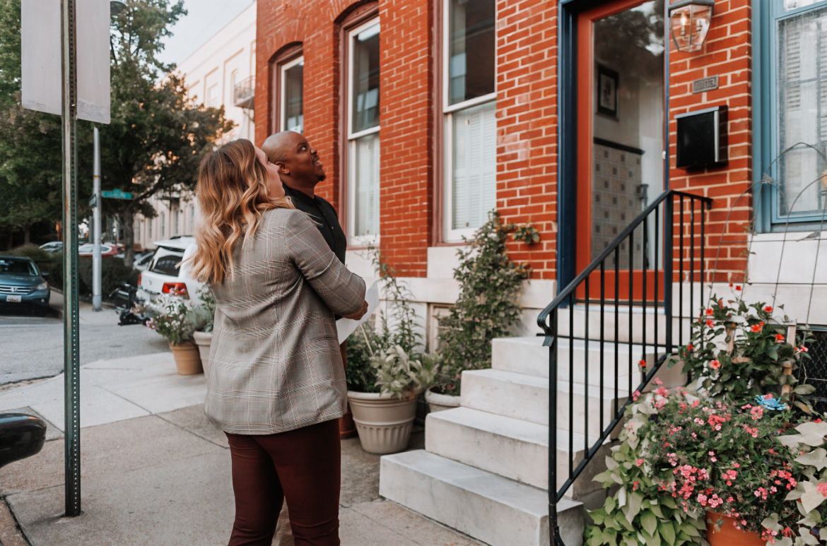 Two women stand on sidewalk near brick building with stairs and potted plants, talking outside a residential entrance.