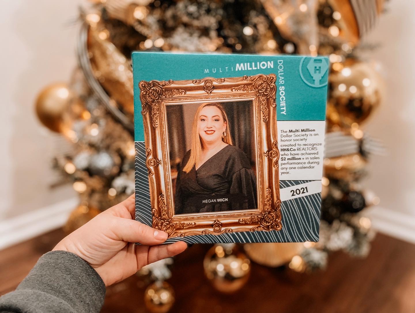 Hand holding a ticket with a woman's photo in a gold frame, Christmas tree in background, gold ornaments, and decorations.