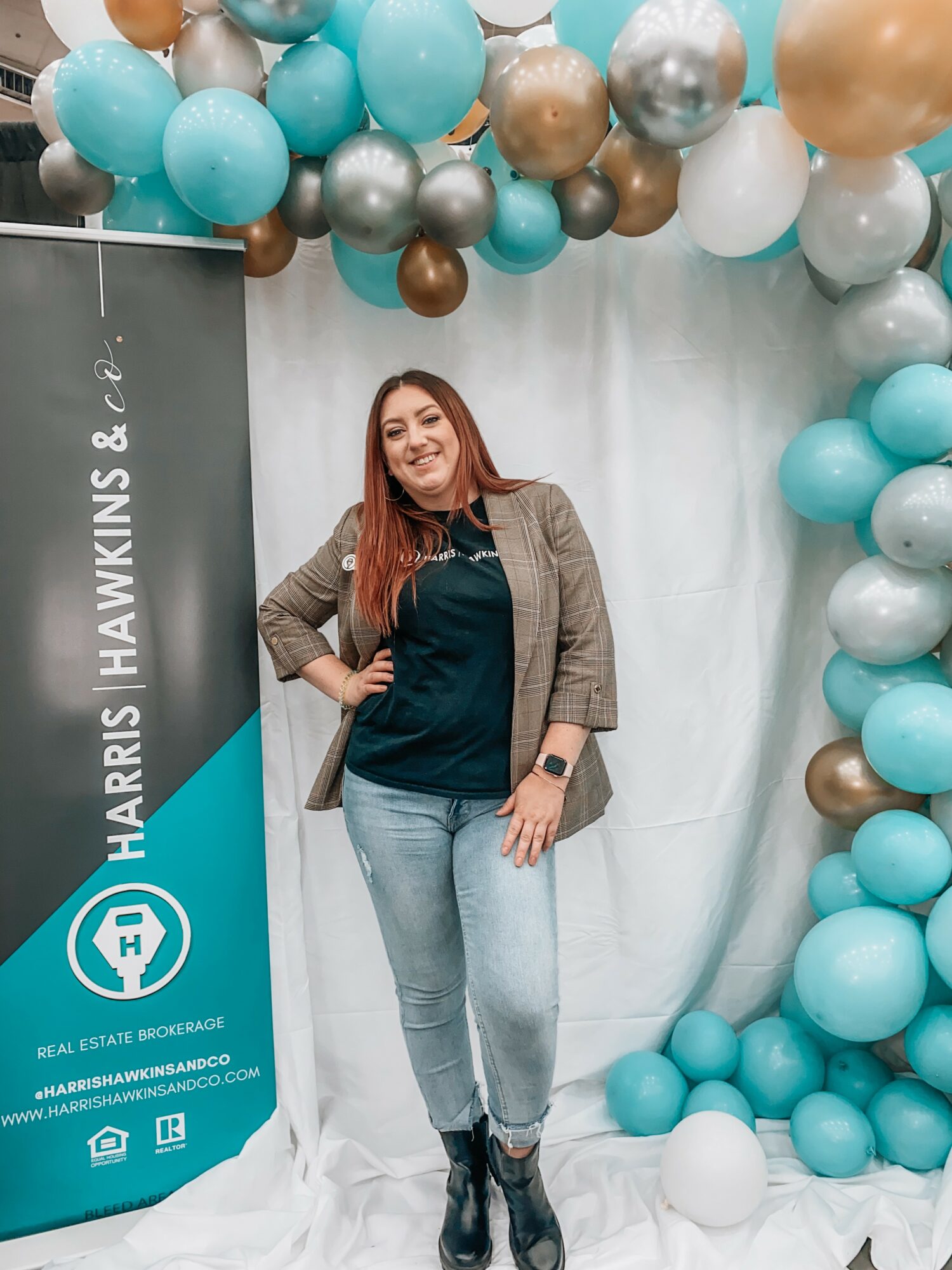 Woman standing in front of a balloon arch, smiling, wearing a blazer, jeans, and boots, with a banner on the side.