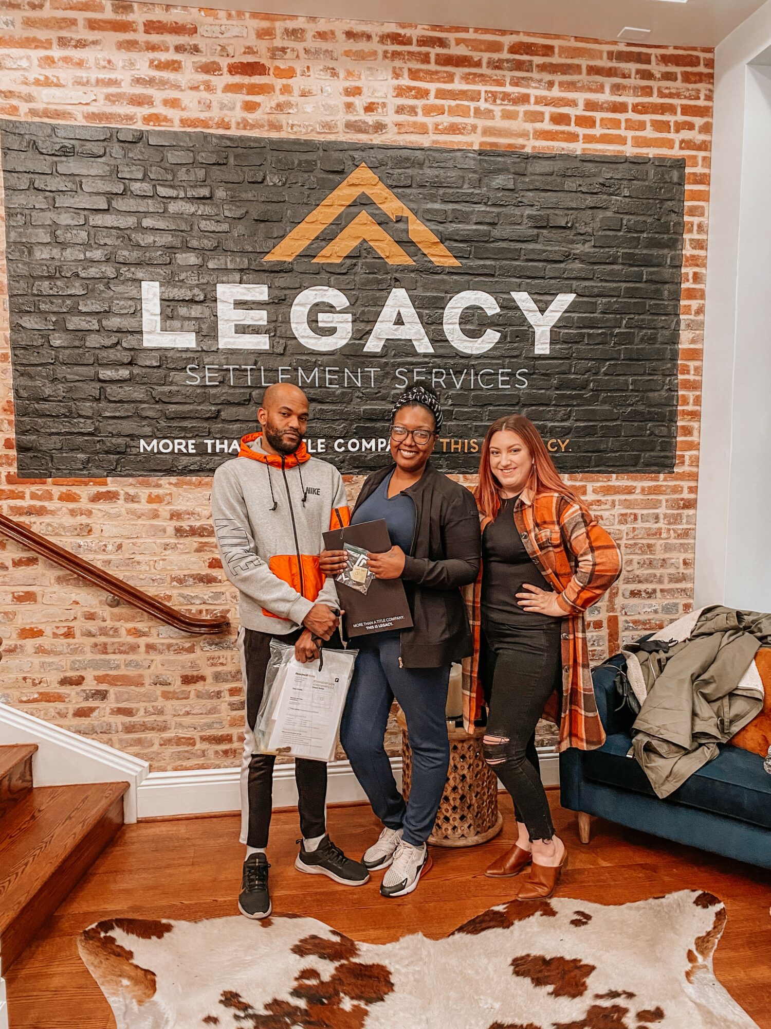 Three people standing in front of a large sign that reads 'LEGACY SETTLEMENT SERVICES' on a brick wall, smiling and holding papers.