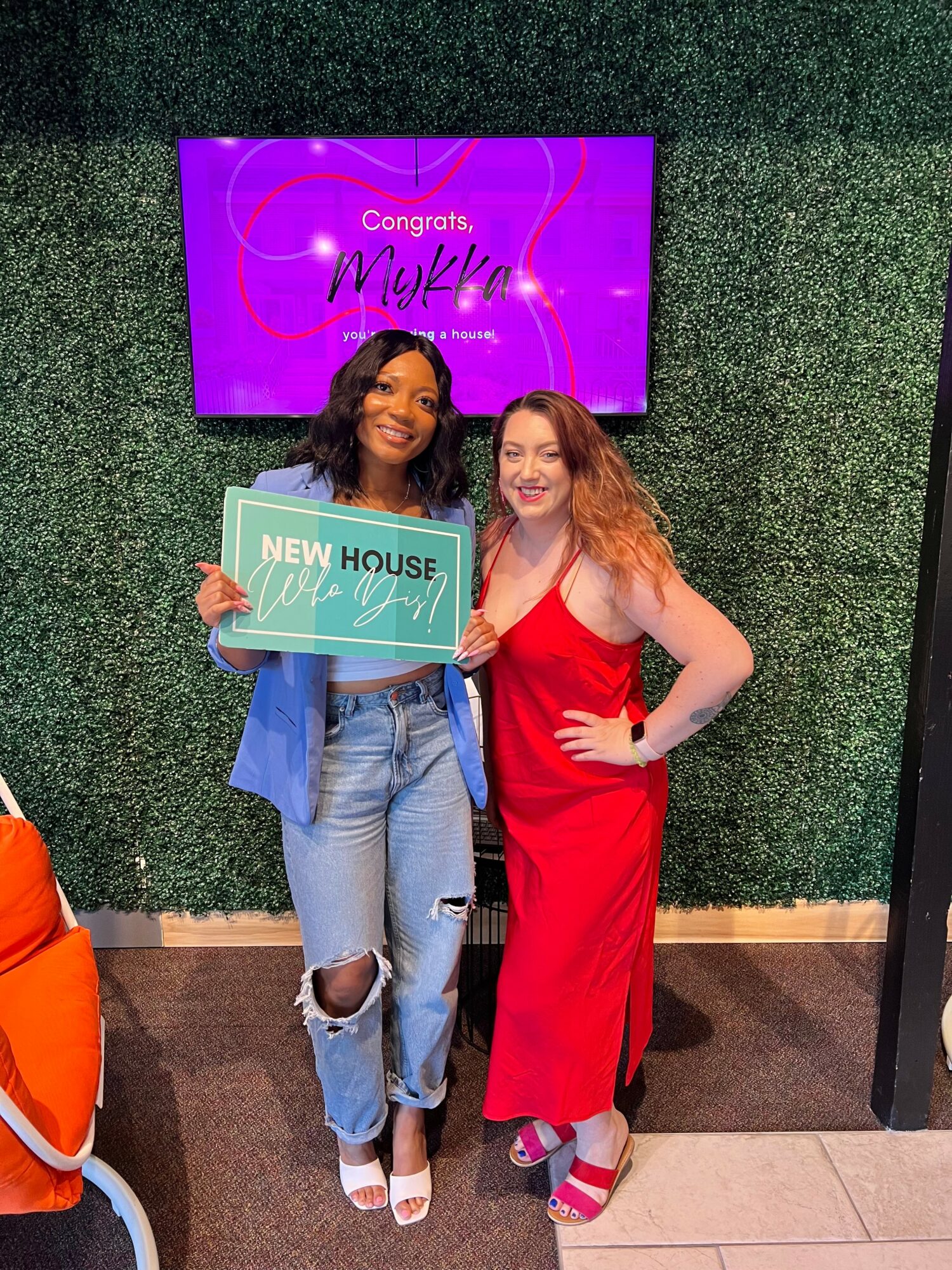 Two women smiling, one holding a sign that says 'New House,' standing in front of a green wall and a screen with congratulatory message.