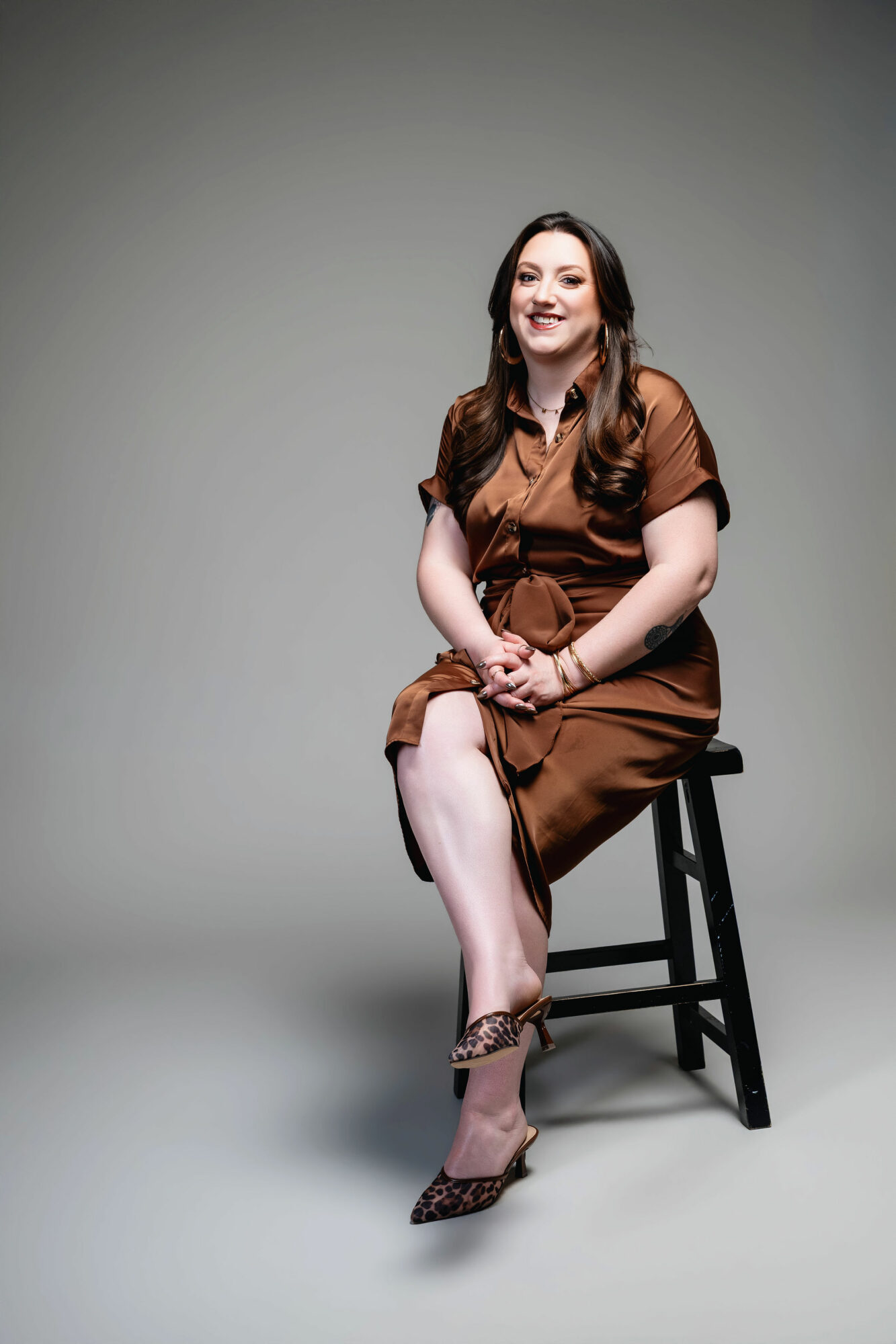 Woman sitting on a stool, smiling, wearing a brown dress and leopard print heels, against a plain background.