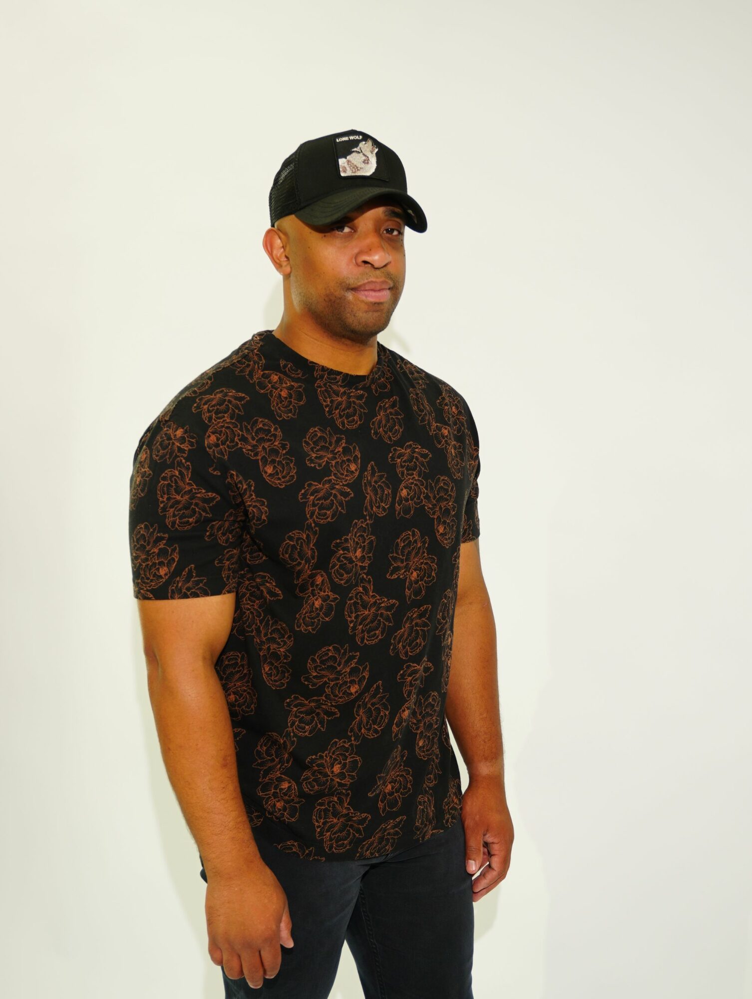 Person standing against a plain white background, wearing a black cap and a patterned shirt, looking to the side.