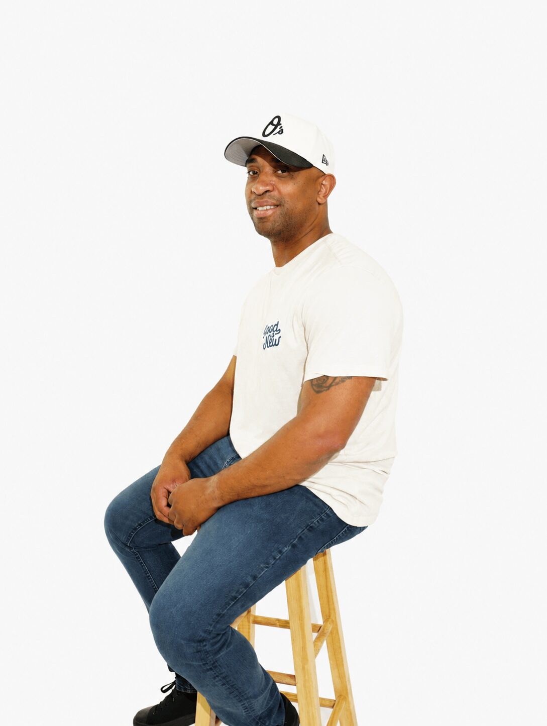 Person sitting on a stool, smiling, wearing a white t-shirt, jeans, and a cap, against a plain background.