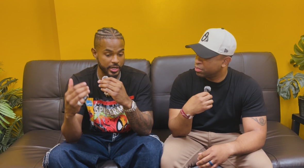 Two men sitting on a black leather couch against a yellow wall, engaged in conversation, with plants on either side.