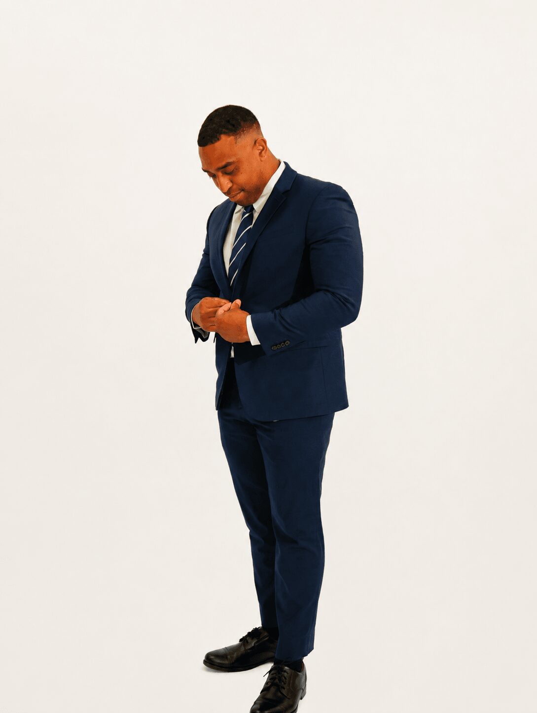 Man in a navy suit adjusting his cuff, standing against a plain white background.