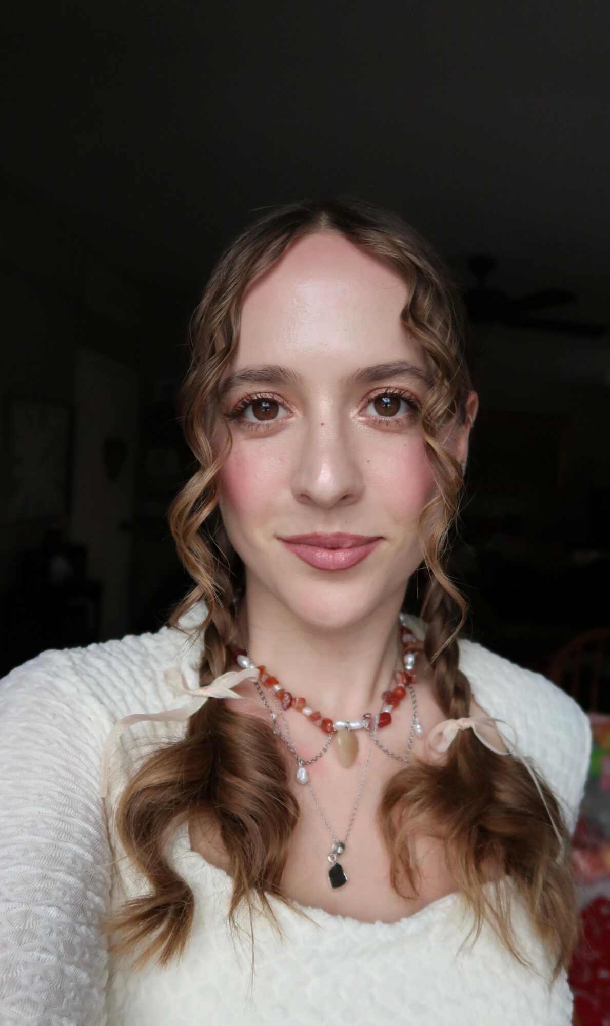 Young woman with wavy hair, wearing a white top and layered necklaces, smiling at the camera.