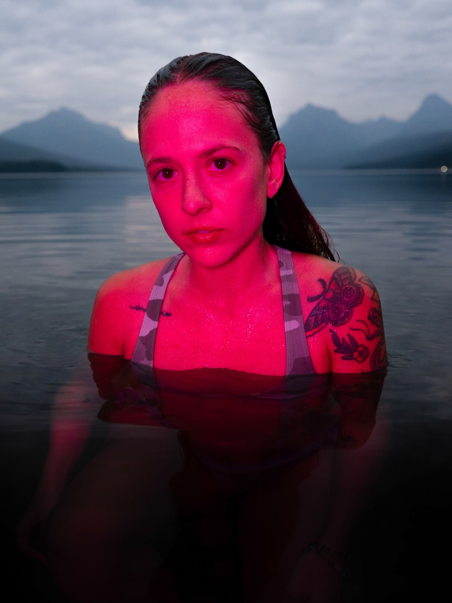 Woman with wet hair and tattoos partially submerged in water, with mountains and cloudy sky in background