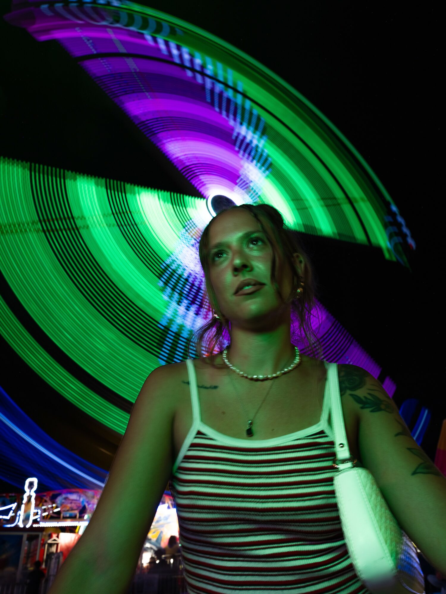 Young woman with striped top and necklace stands in front of colorful light display at night.