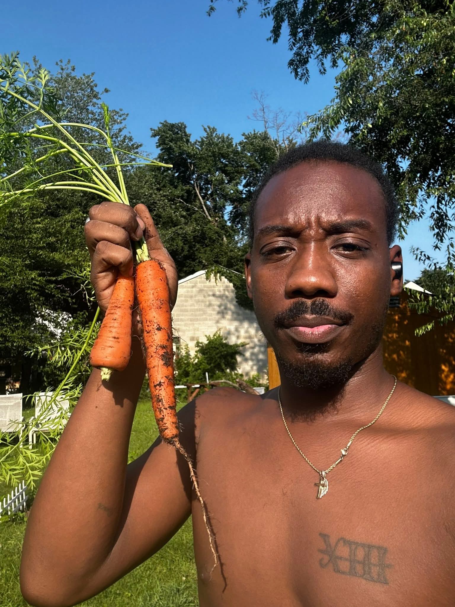 Man holding two carrots outdoors with trees and blue sky in background.